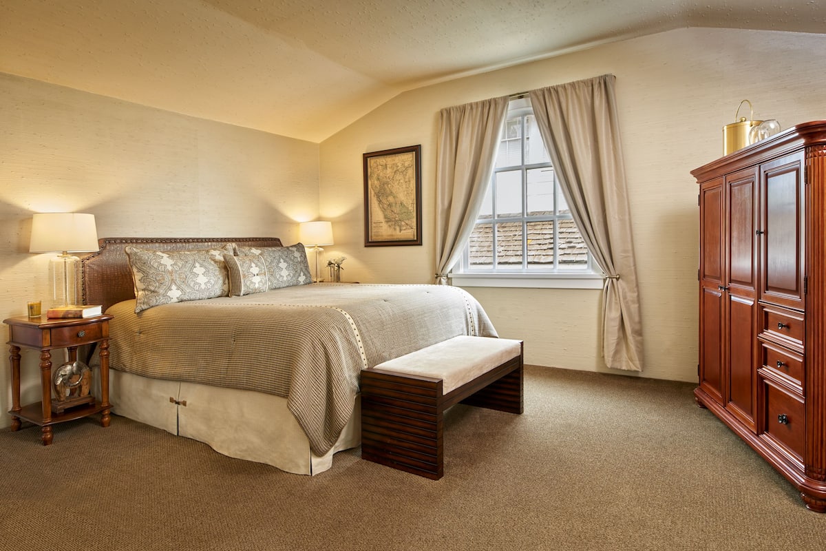 A bedroom with a large bed, bedside lamps, a wooden nightstand, a bench, a wardrobe, and a window with curtains.