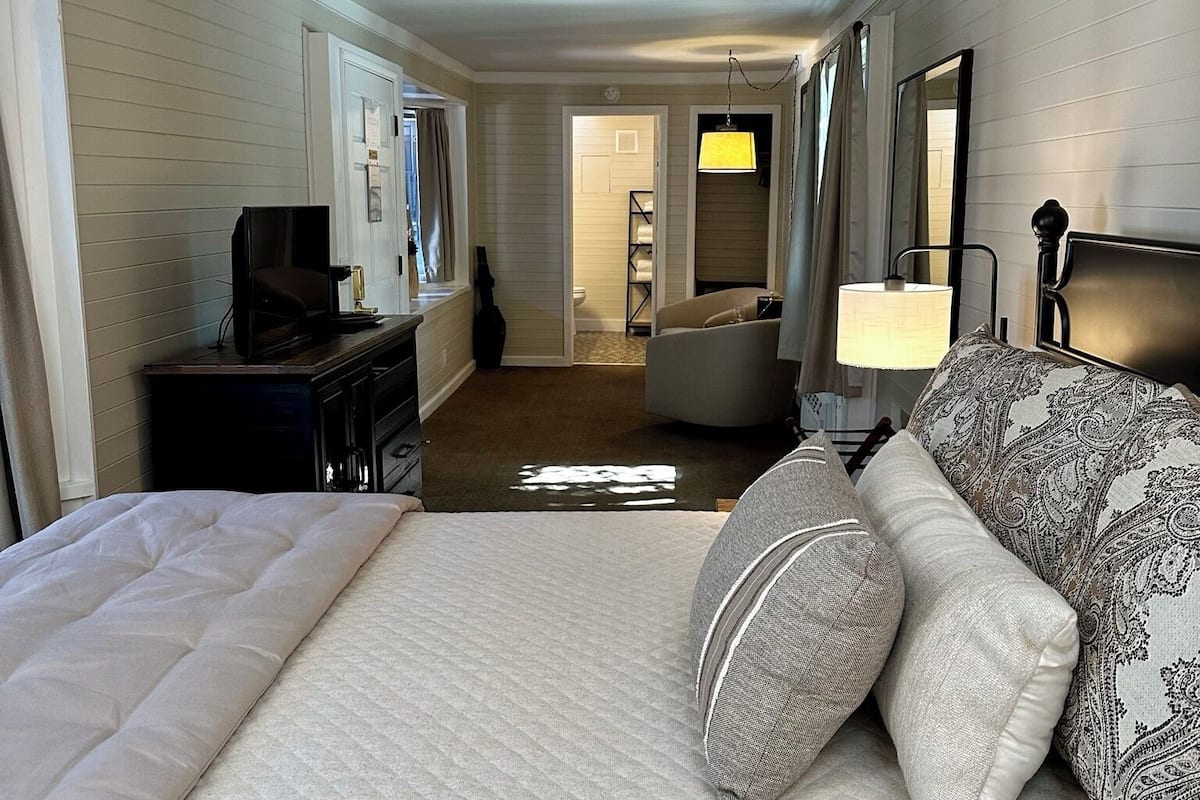 A hotel room with a bed, a sofa, a desk with a television, a mirror, and a bathroom visible in the background.