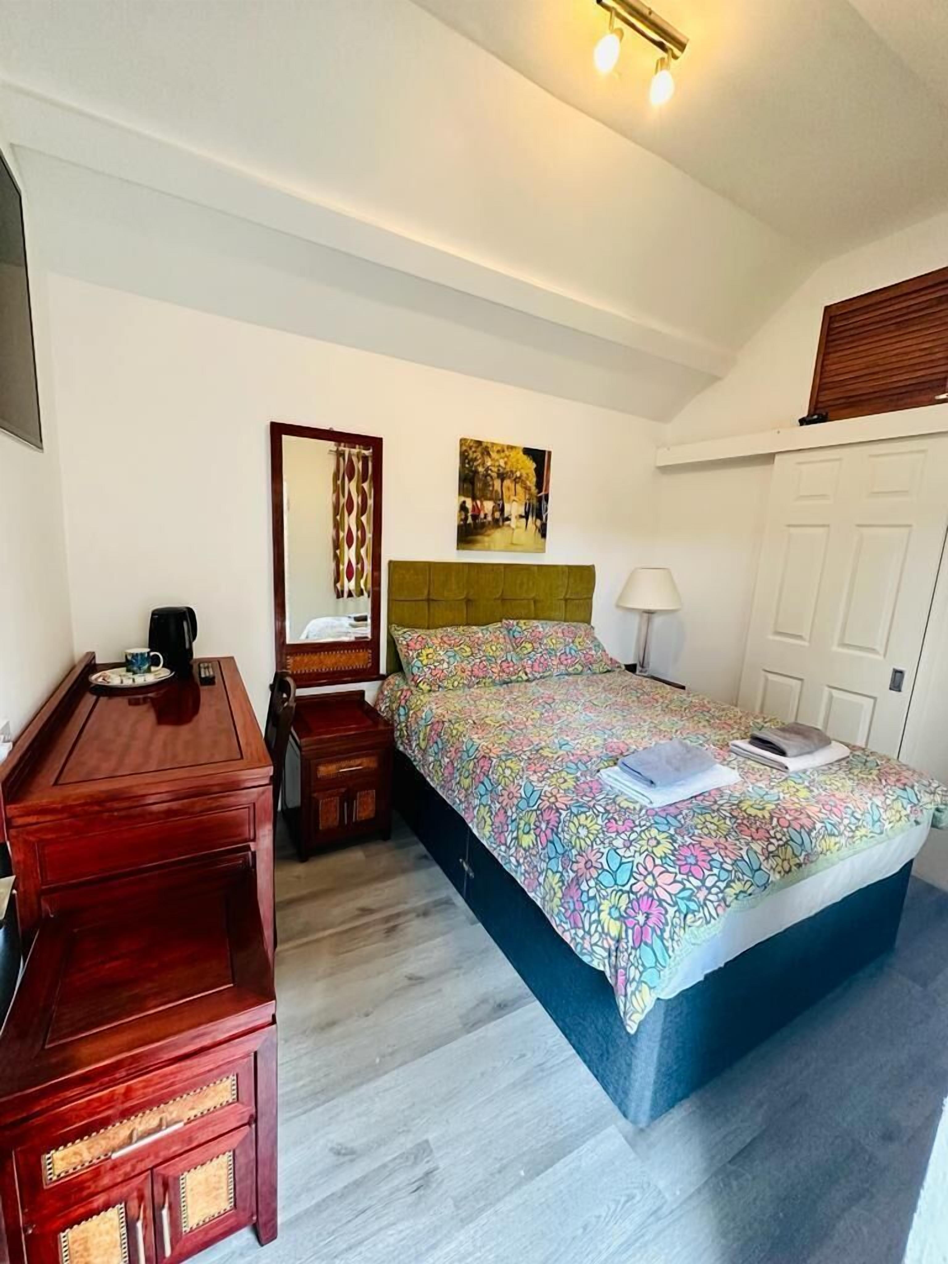 Un dormitorio con una cama, un aparador de madera, una mesita de noche, una lámpara, un espejo y una puerta.