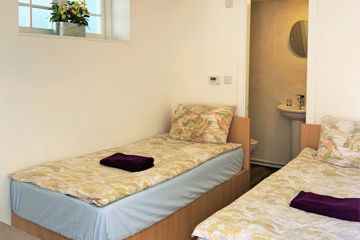A small, clean room with two single beds, a window with a plant, and a bathroom visible through an open door.