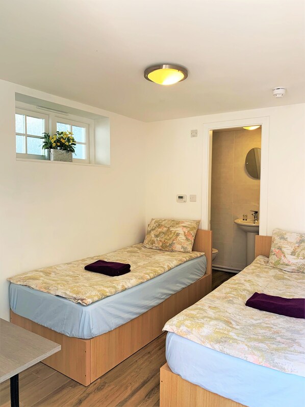A small, clean room with two single beds, a window with a plant, and a bathroom visible through an open door.