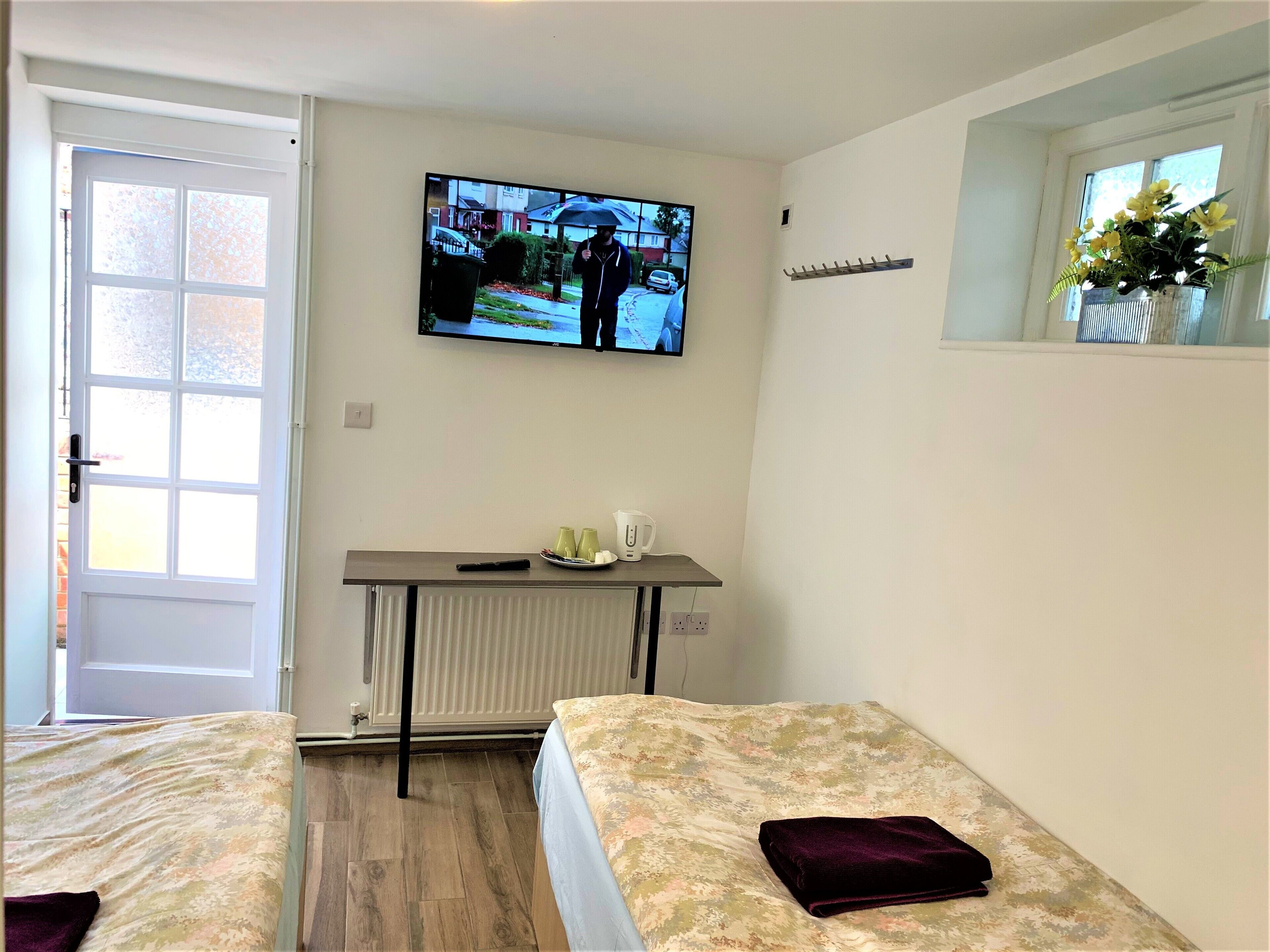 A room with two beds, a small table with a kettle and cups, a radiator, and a TV on the wall.