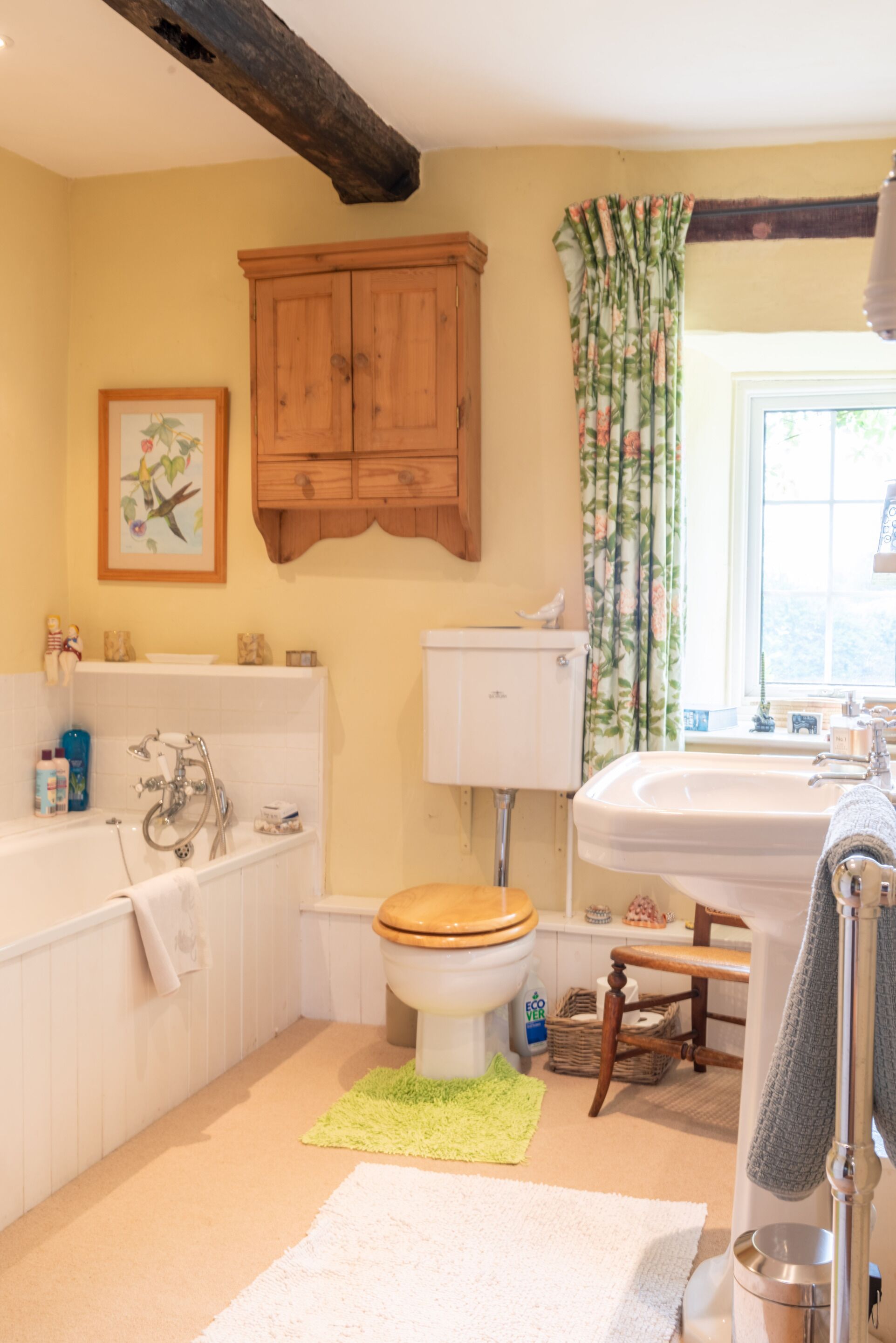 Ein Badezimmer mit Badewanne, Toilette und Waschbecken. Über der Toilette befindet sich ein Holzschrank und an der Wand ein gerahmtes Bild.