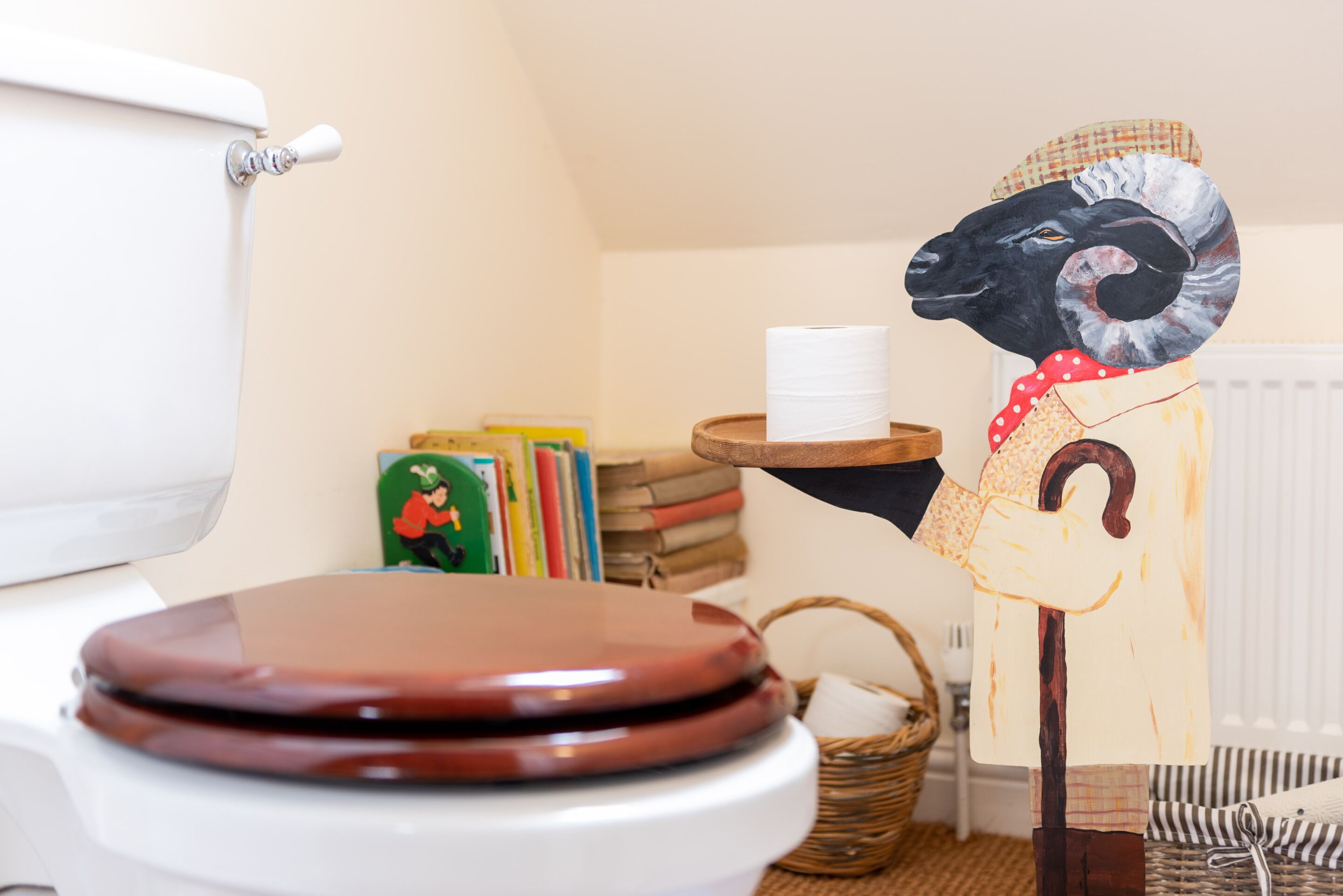 A toilet with a brown seat, a roll of toilet paper on a wooden holder, and a wall-mounted shelf with books and a basket.