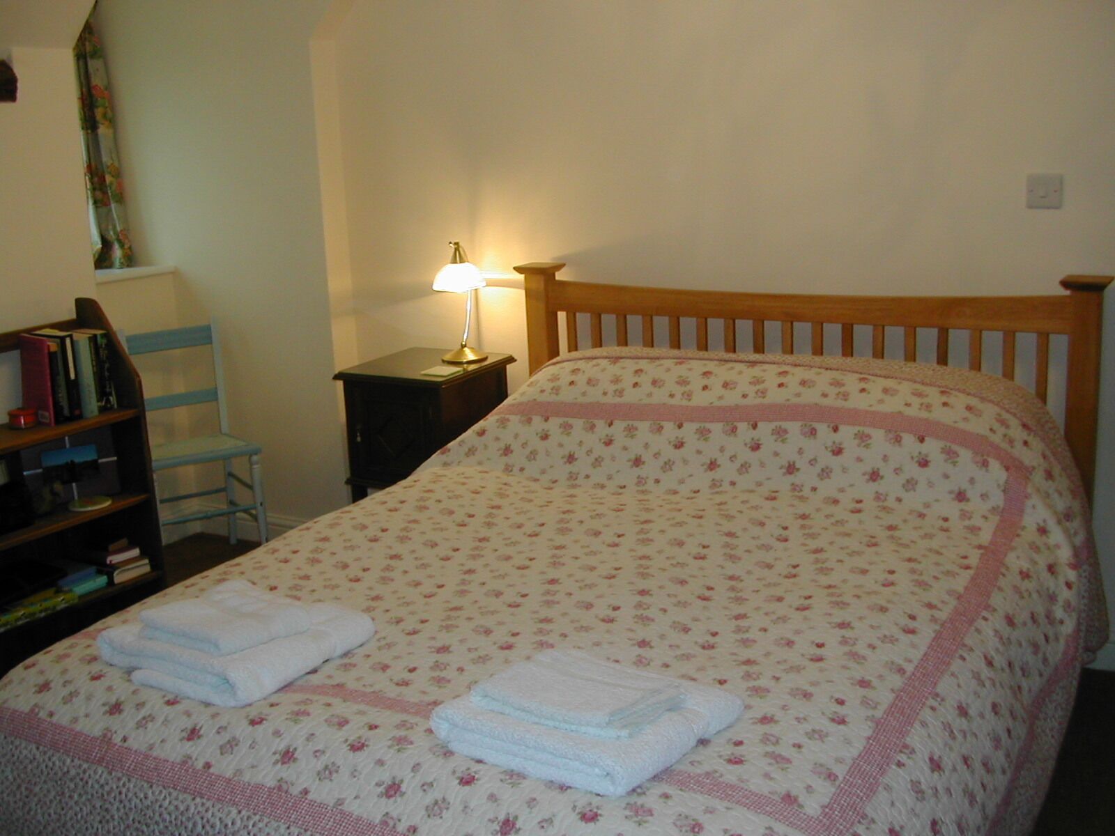 A bedroom with a wooden bed, a nightstand with a lamp, a chair, and a bookshelf.