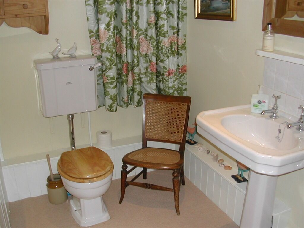 A bathroom with a toilet, wooden chair, sink, and floral curtains.