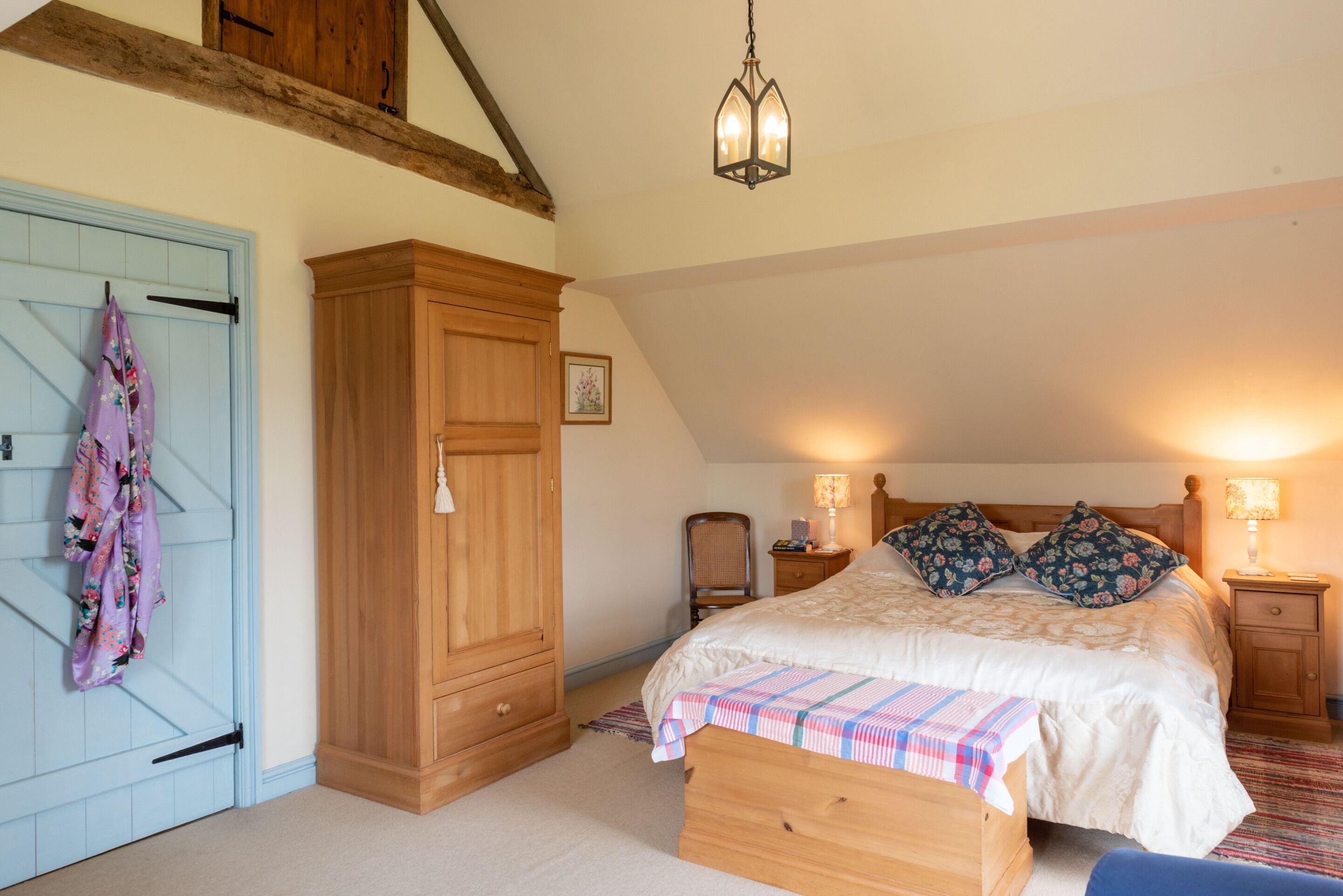 A bedroom with a large bed, a wooden wardrobe, a hanging light fixture, and a nightstand with a lamp.