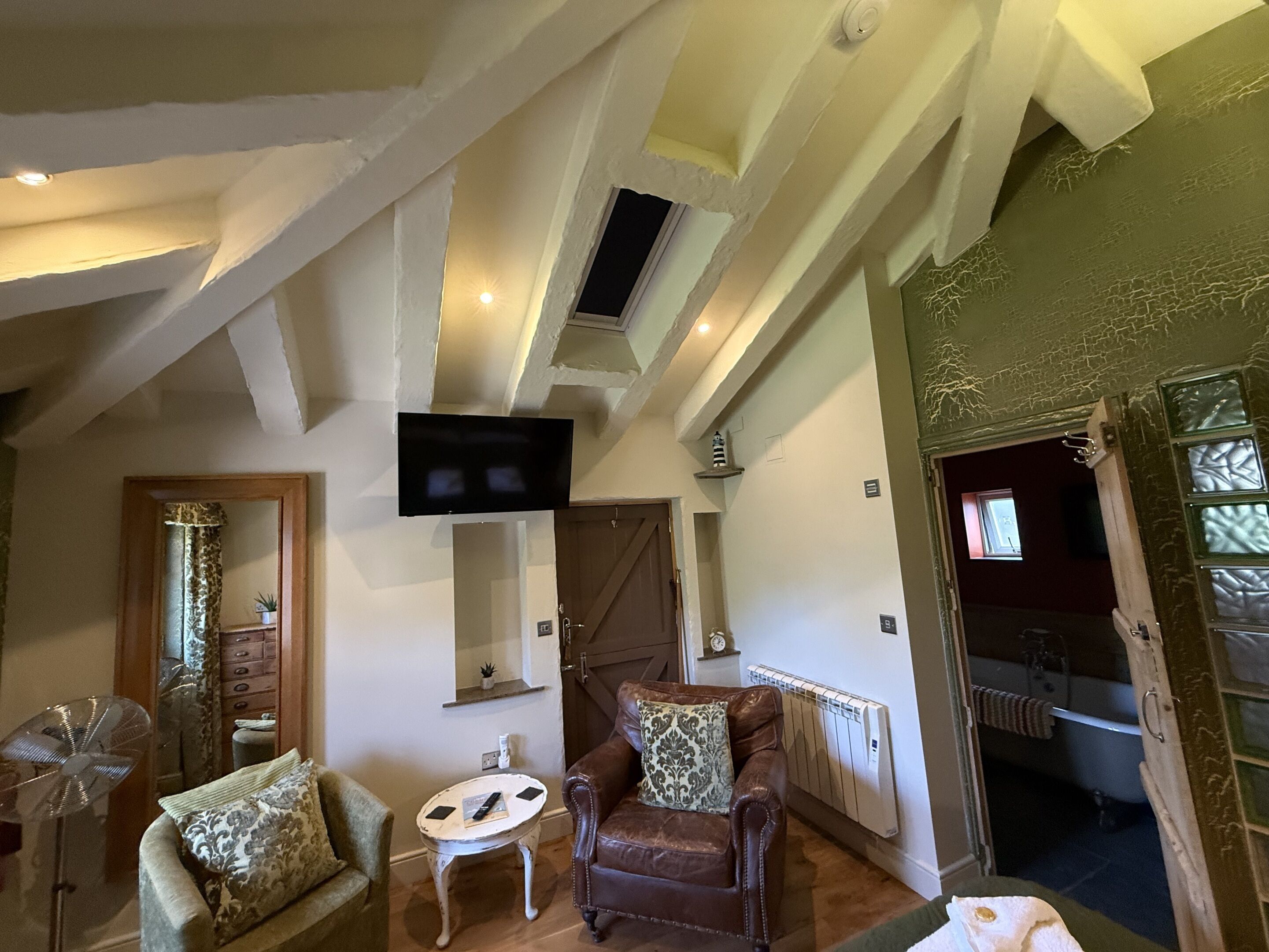 A room with a brown leather sofa, a small round table, a television mounted on the wall, and a window with a view of the outside.