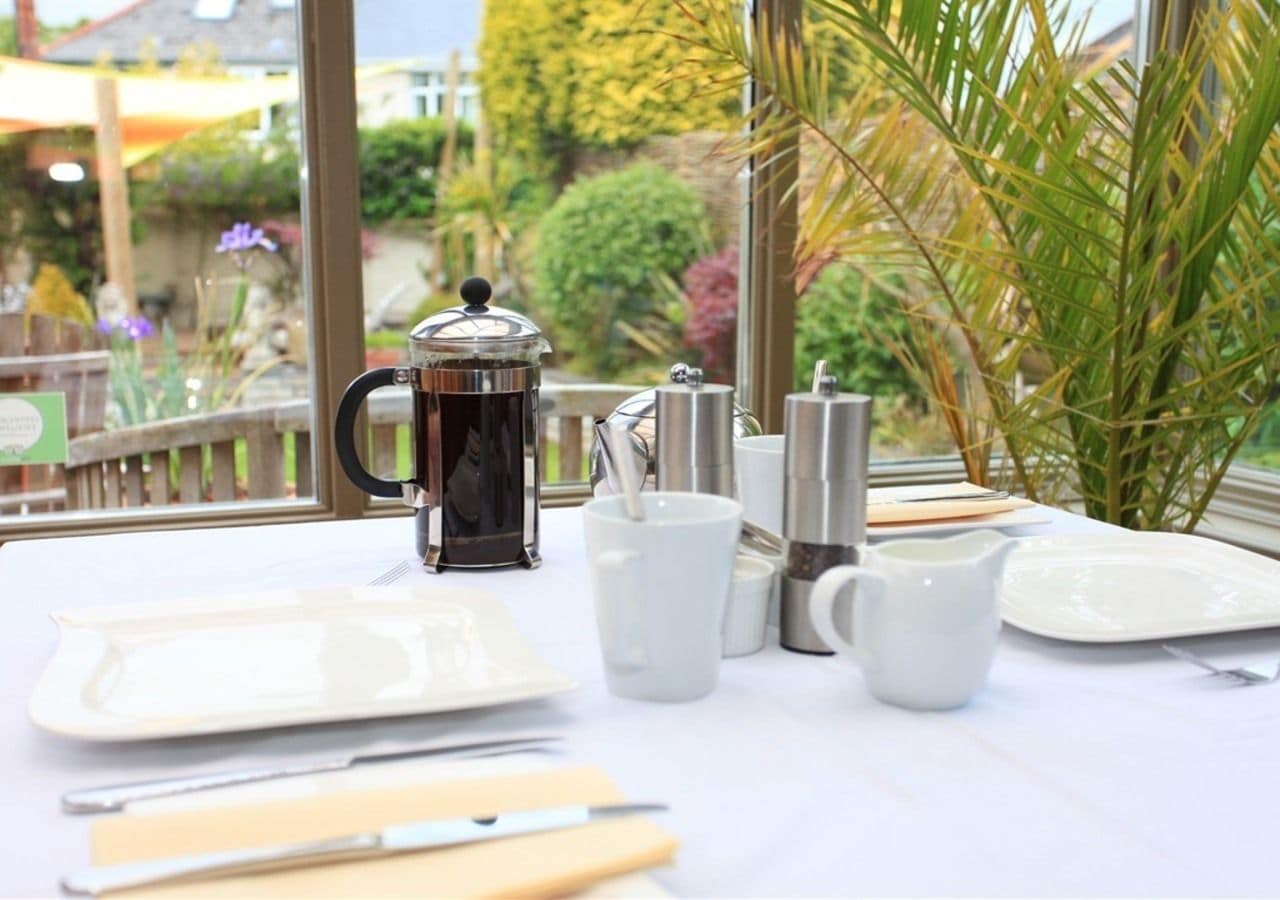 Una mesa servida con una cafetera, tazas, platos y cubiertos, con vistas a la vegetación y un edificio en el exterior.