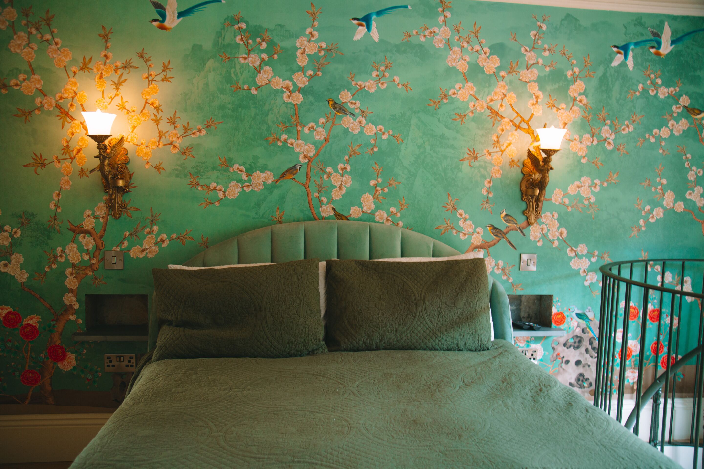 A bedroom with a green upholstered headboard, two wall-mounted lamps, and a mural of birds and flowers on the wall.