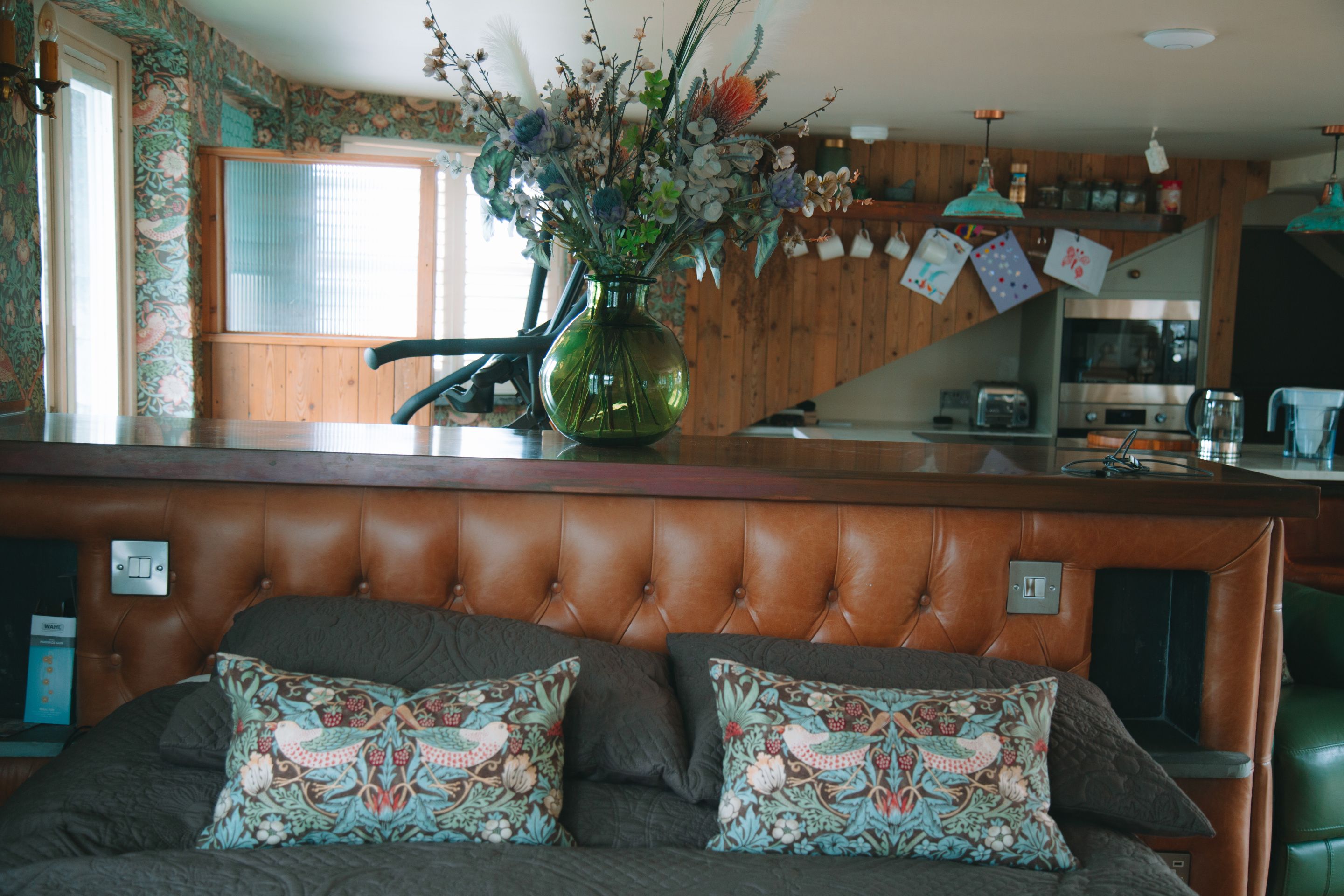 A room with a tufted headboard, a bed with patterned pillows, a green vase with flowers, and a kitchen area in the background.