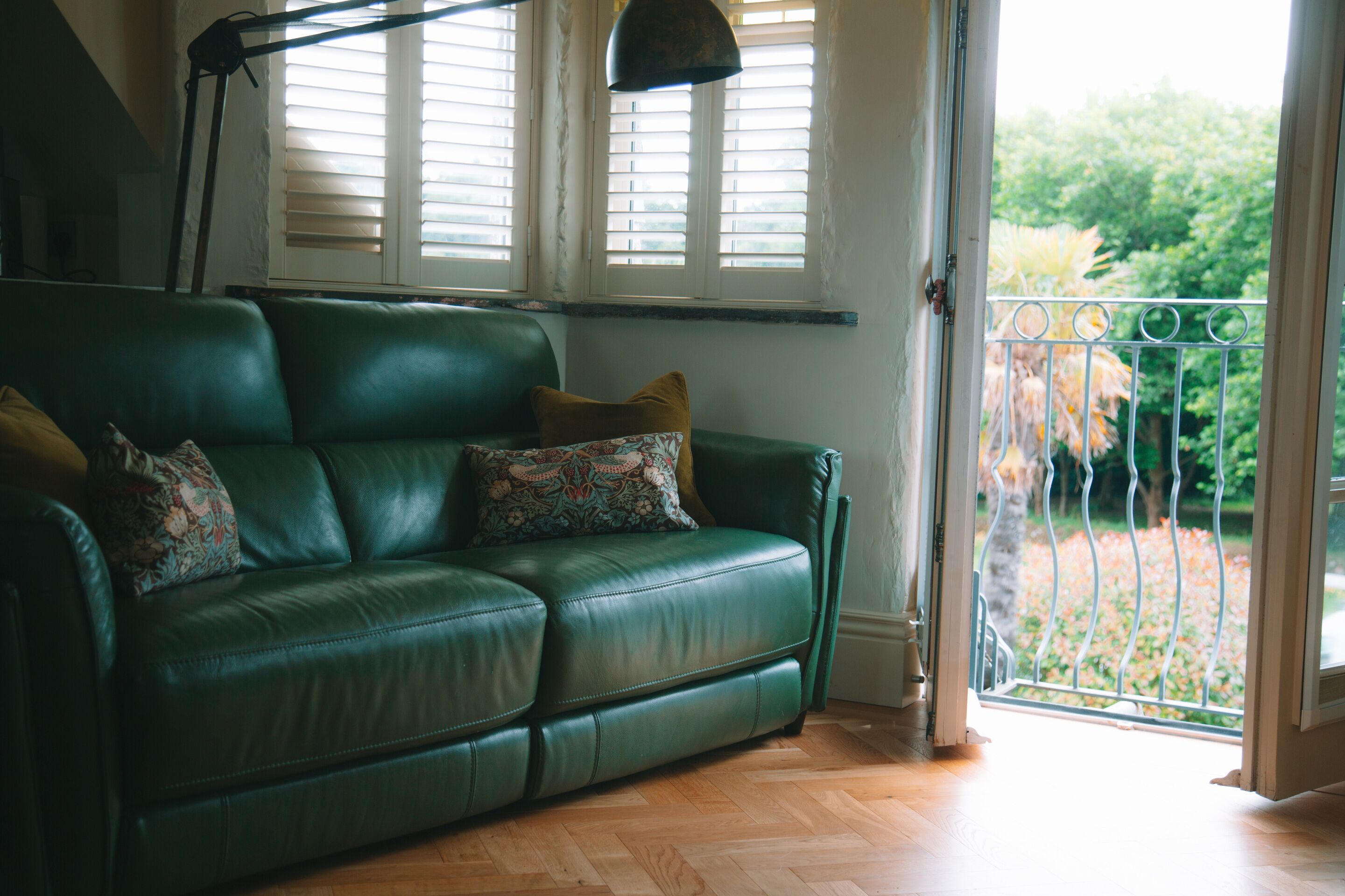 A green leather sofa with patterned cushions, a floor lamp, and a view of a garden through a balcony door.