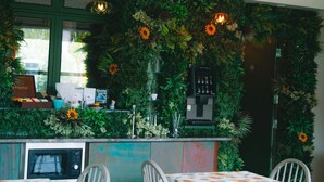 Restaurant amb una paret verda decorada amb plantes i flors, una màquina de cafè i taules amb cam amesa floral.