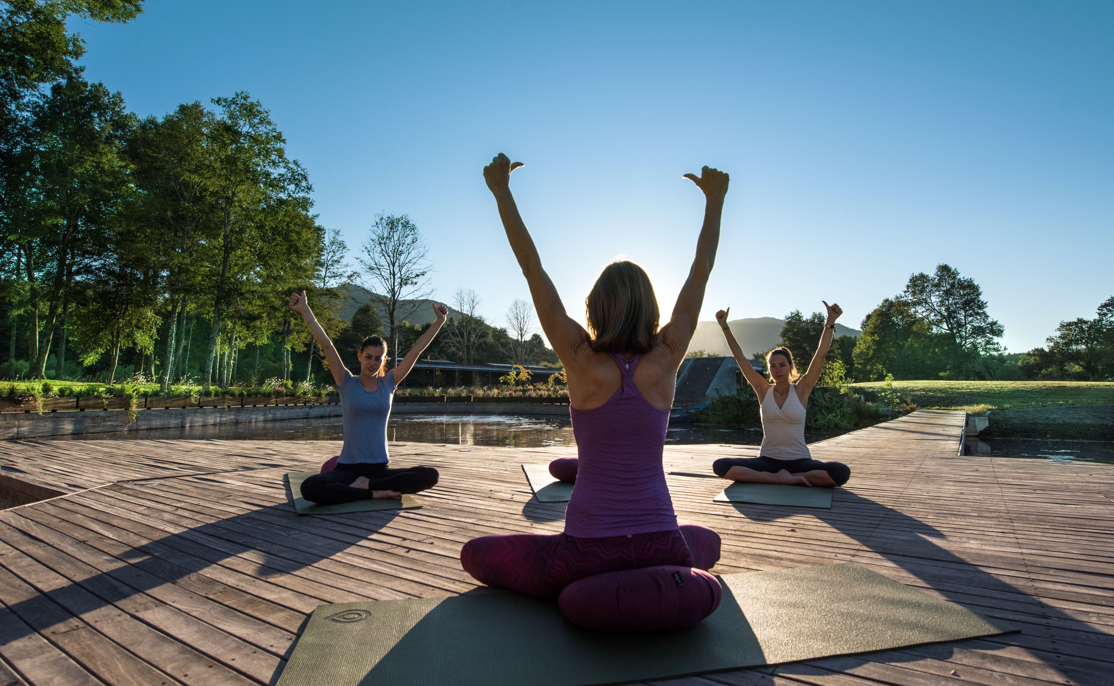 Tres personas practicando yoga en una terraza de madera con un lago y árboles al fondo.