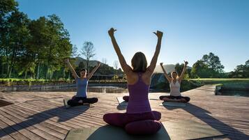 Tres personas practicando yoga en una terraza de madera con un lago y árboles al fondo.