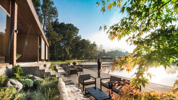 Una terraza de madera con asientos al aire libre, vista a un lago y un edificio con ventanas amplias.