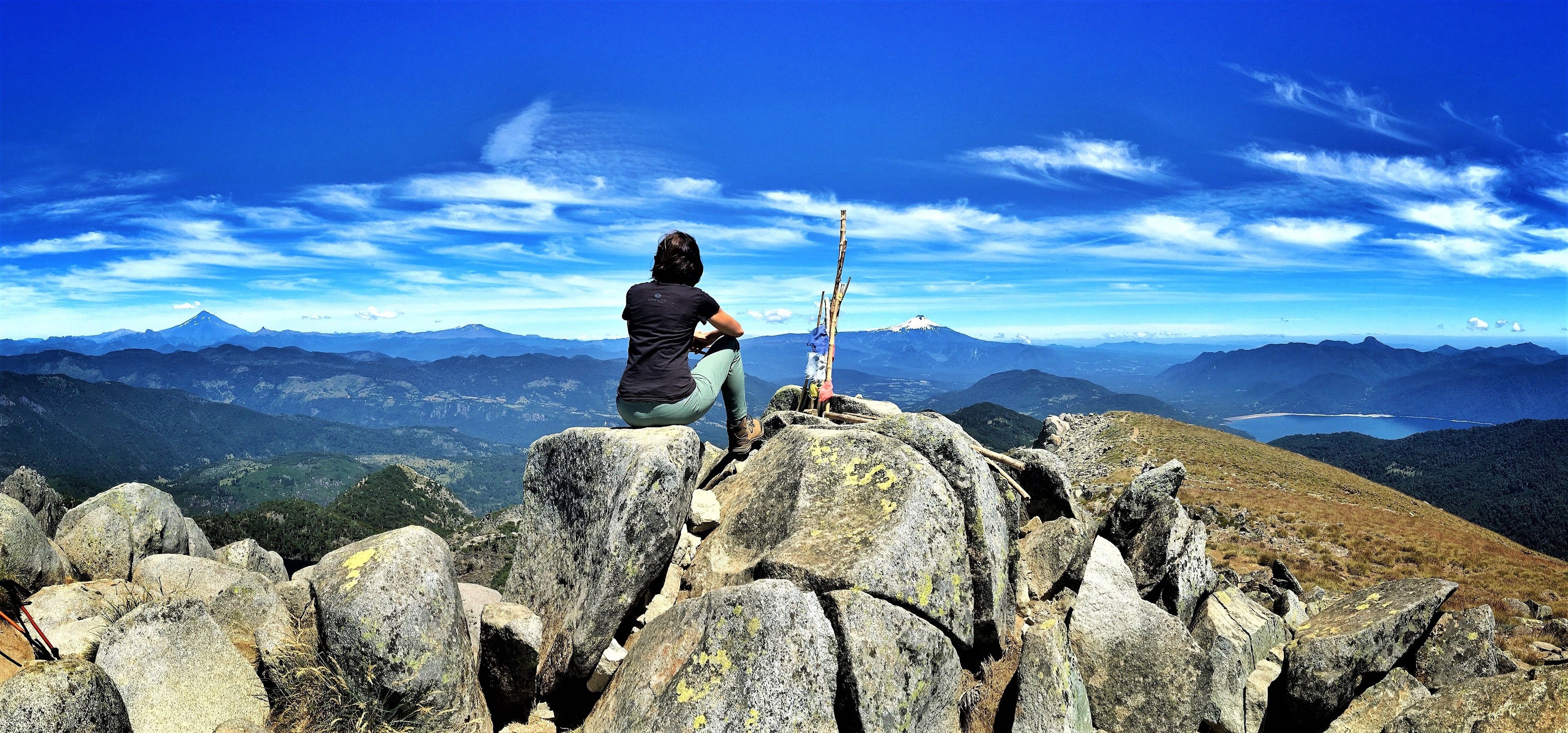 Una persona sentada sobre un saliente rocoso con vista a un paisaje montañoso bajo un cielo despejado.