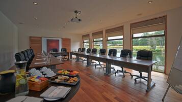 A conference room with a long table, chairs, and a projector. There are windows with a view of greenery outside.