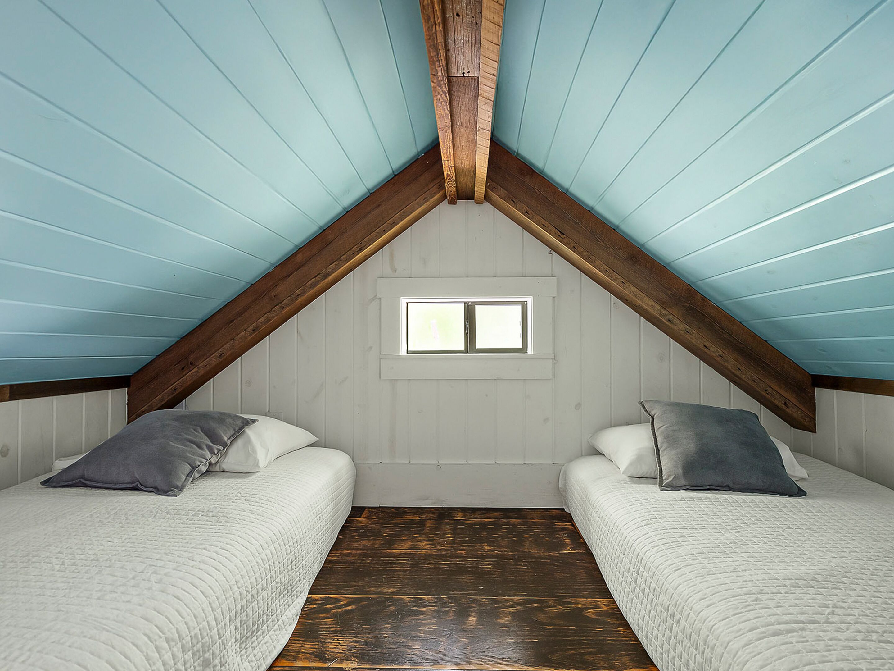 A room with a sloped ceiling, wooden beams, two beds with pillows, a small window, and a wooden floor.
