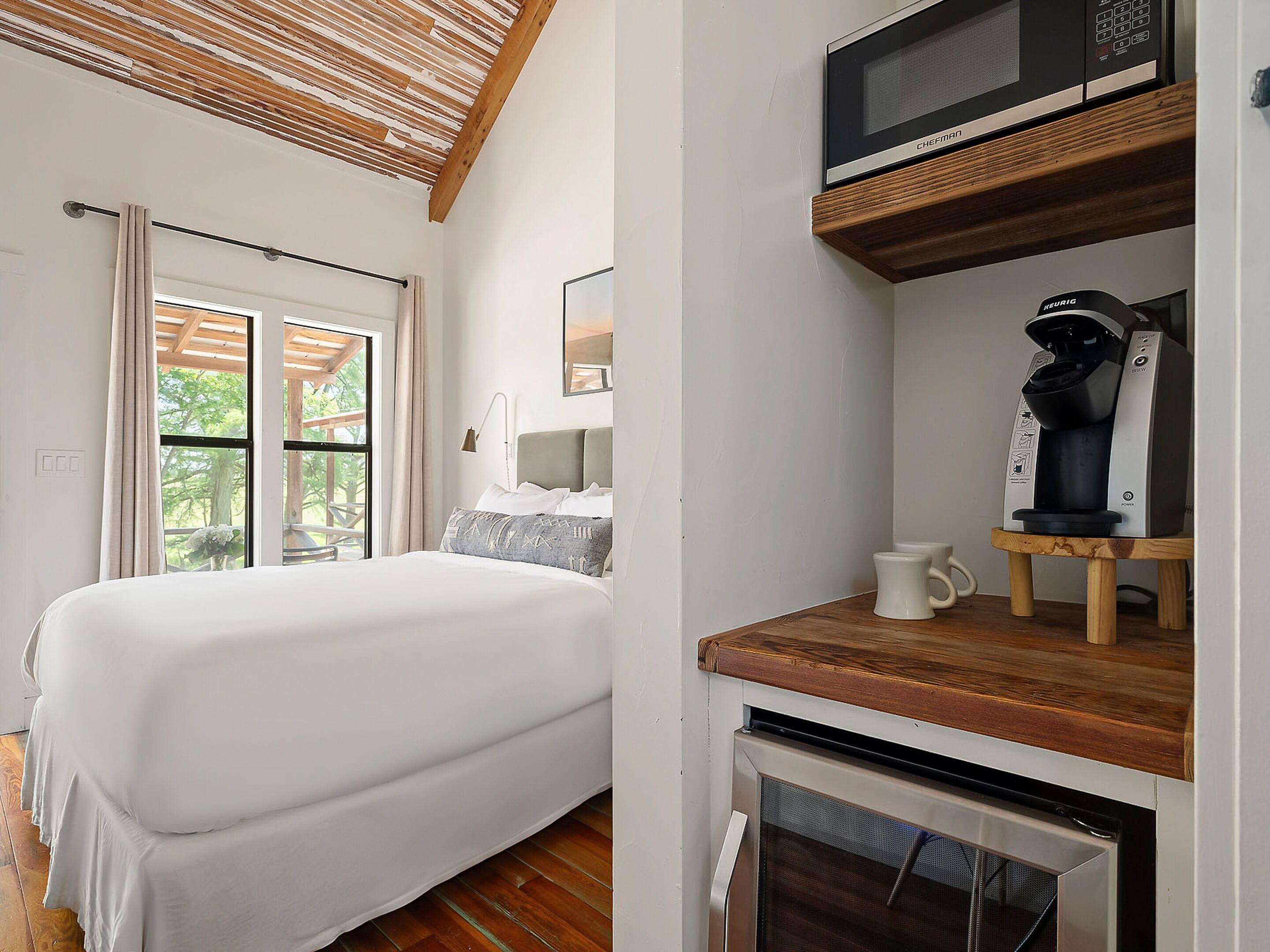 A bedroom with a bed, a window, a ceiling with wooden beams, a microwave, and a coffee maker.