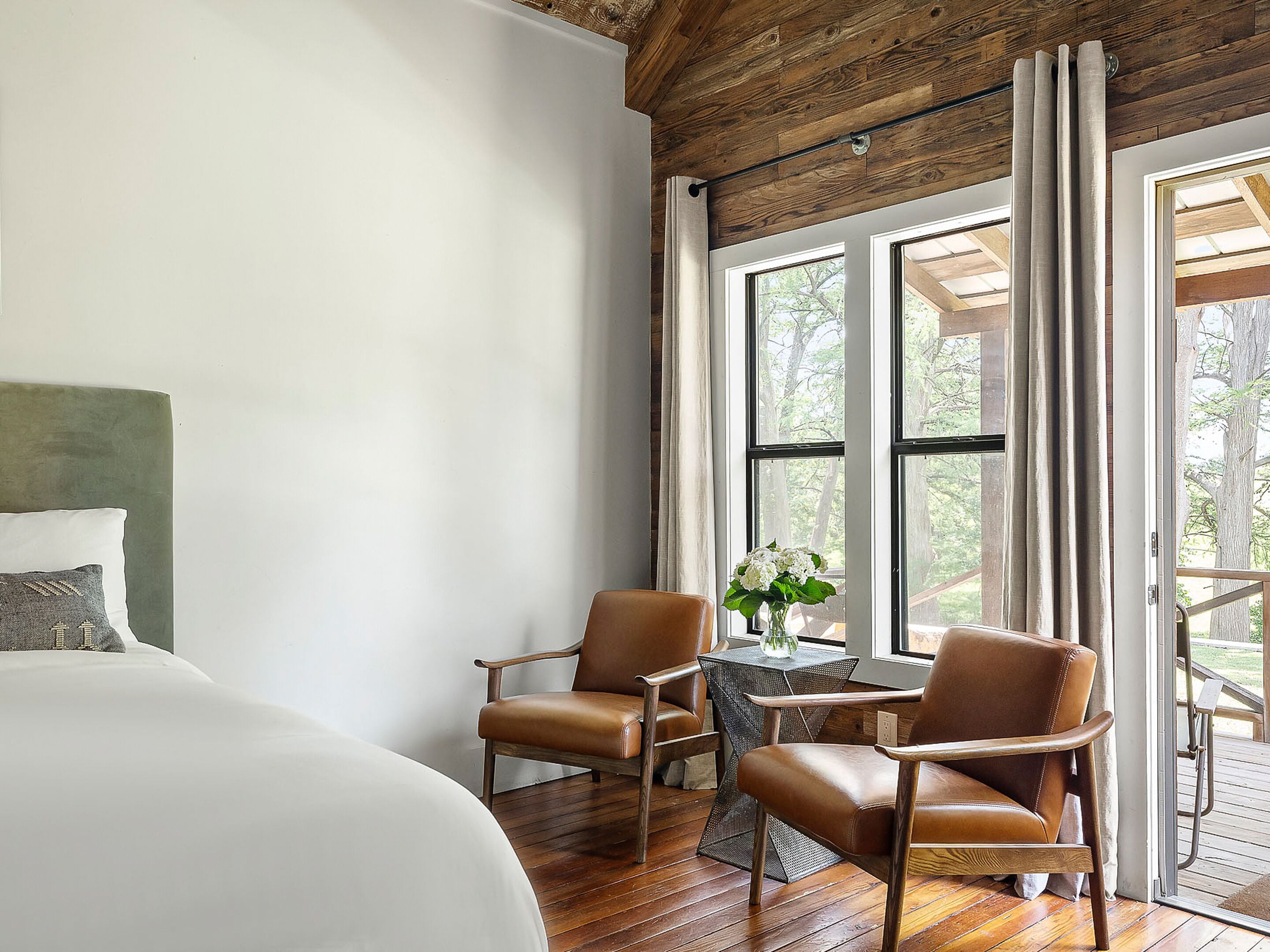 A bedroom with a large bed, two leather chairs, a small table with a vase of flowers, and large windows with curtains.