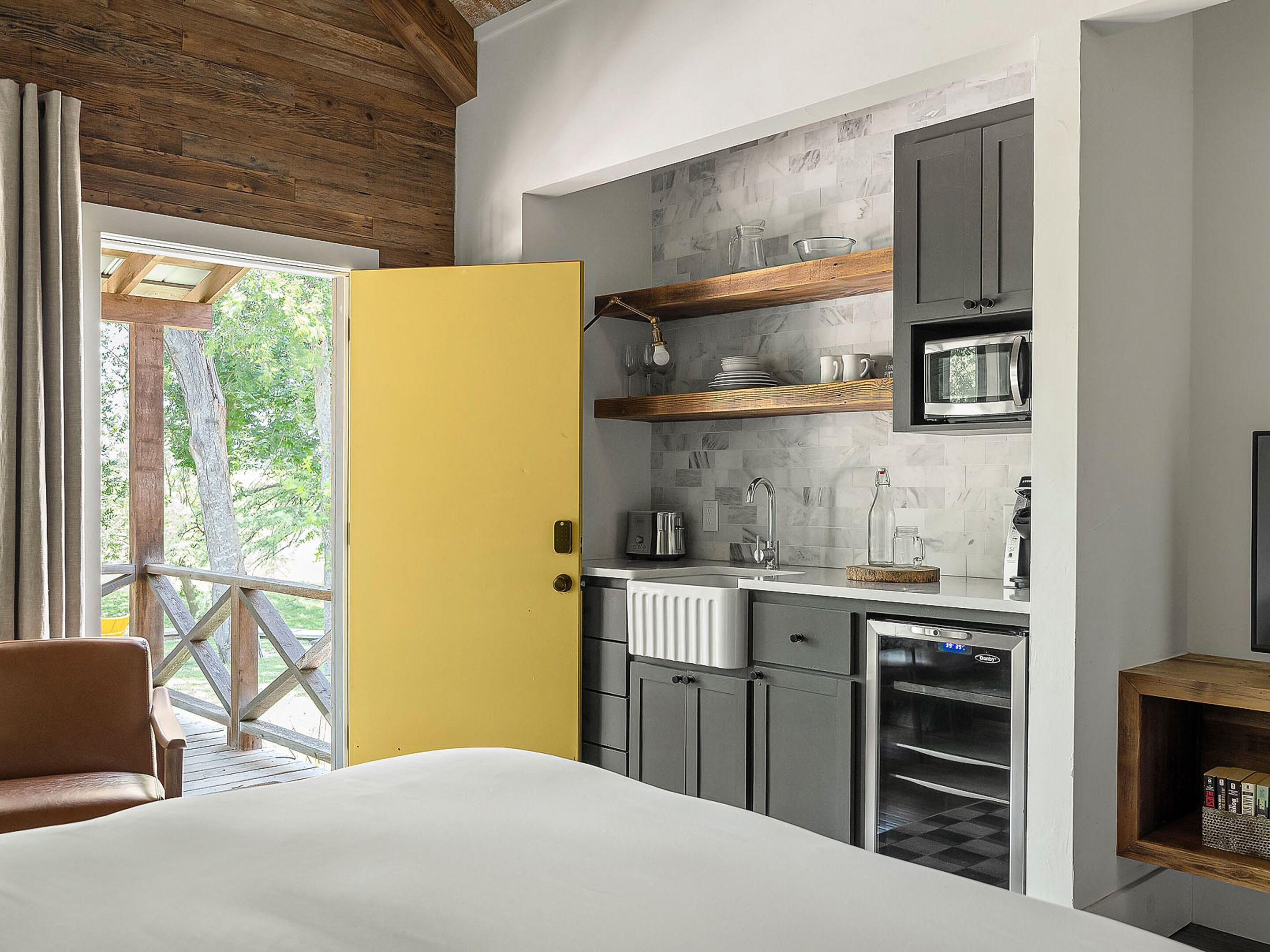 A compact kitchen with a yellow door, wooden ceiling, and a view of greenery outside.