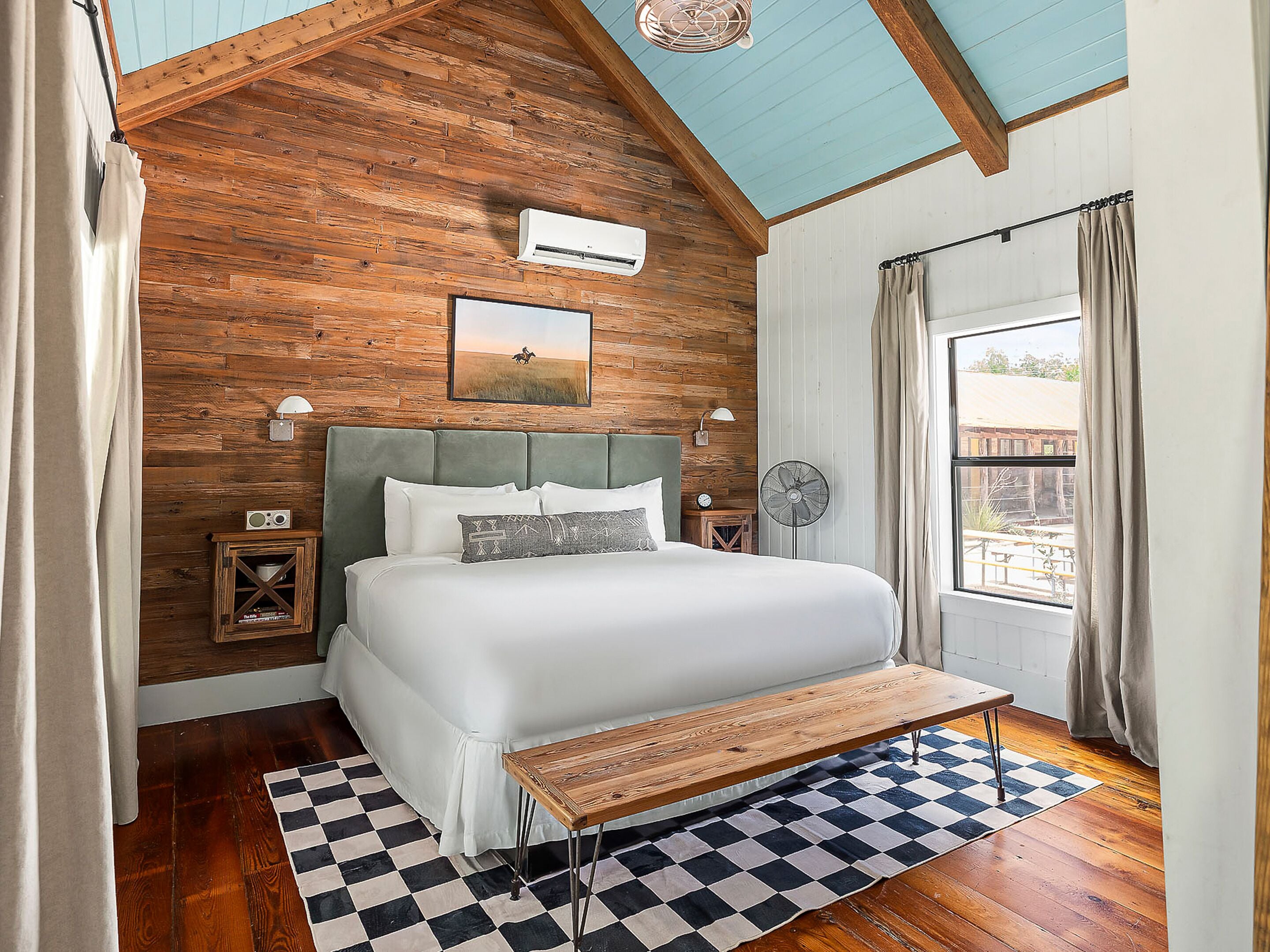 Une chambre avec un plafond en bois, un lit recouvert de draps blancs, un tapis à carreaux et un banc en bois.