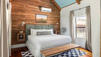 Une chambre avec un plafond en bois, un lit recouvert de draps blancs, un tapis à carreaux et un banc en bois.