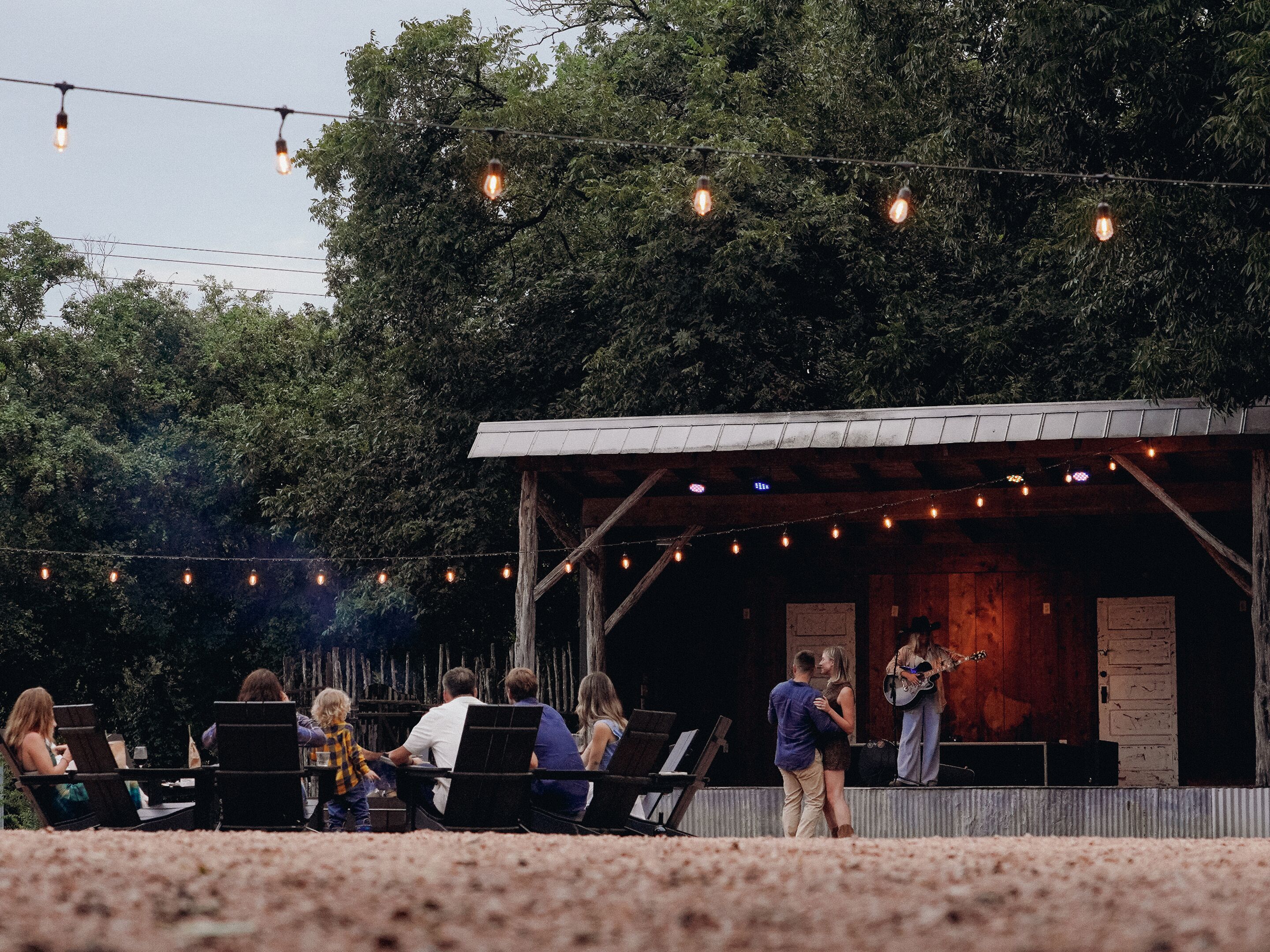 An outdoor seating area with string lights, a stage, and a rustic wooden structure.