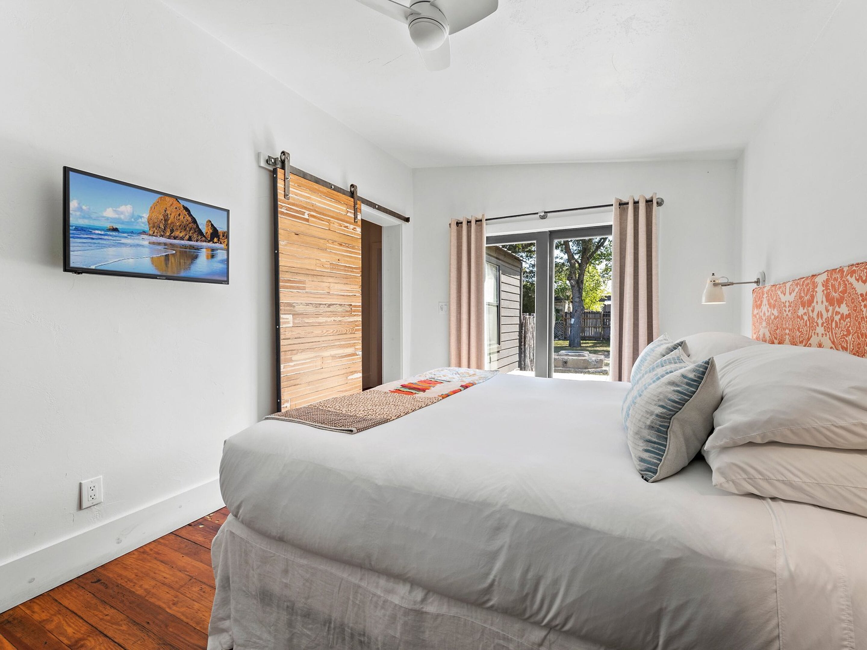 A bedroom with a bed, a view of the outdoors, a ceiling fan, and a painting on the wall.