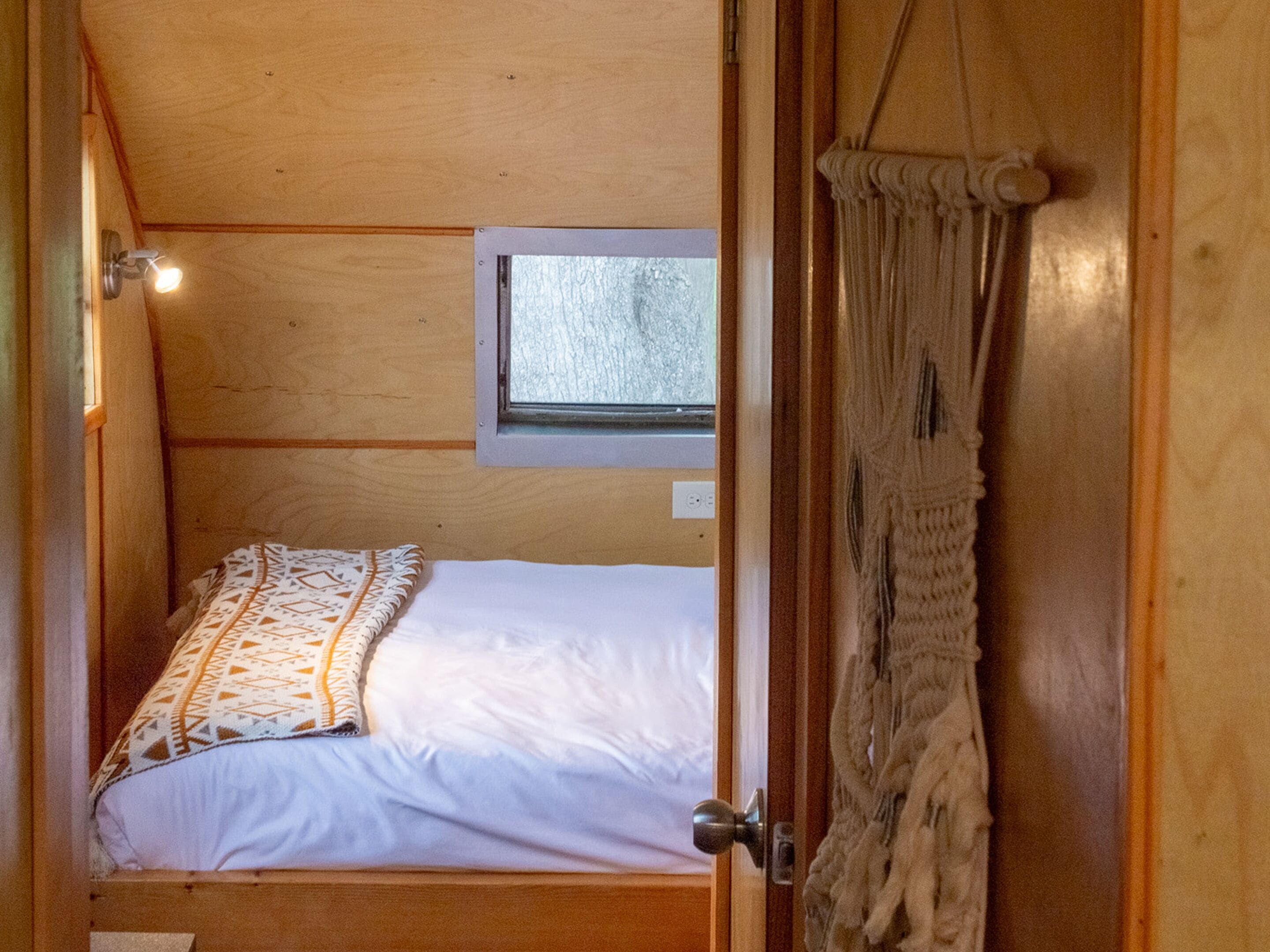 A compact bedroom with a single bed, a small window, and a macramé curtain.