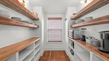 A narrow kitchen with wooden shelves and white walls, featuring a window with blinds.