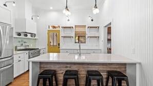 Uma cozinha moderna com armários brancos, uma ilha grande com banquetas, eletrodomésticos de aço inoxidável e iluminação pendente.