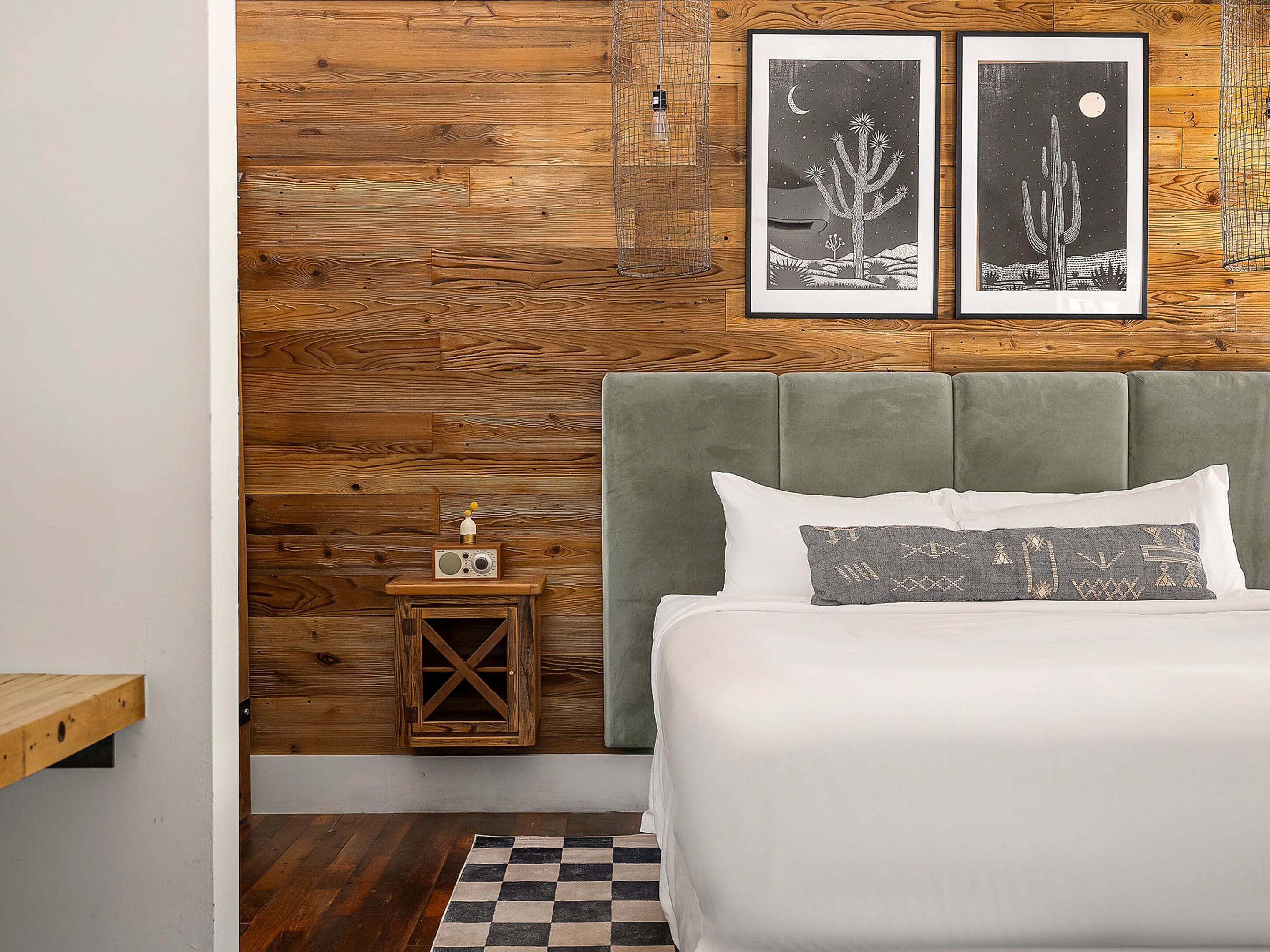 A bedroom with a wooden wall, a bed with white bedding, a small wooden nightstand with a radio, and two framed pictures of cacti.