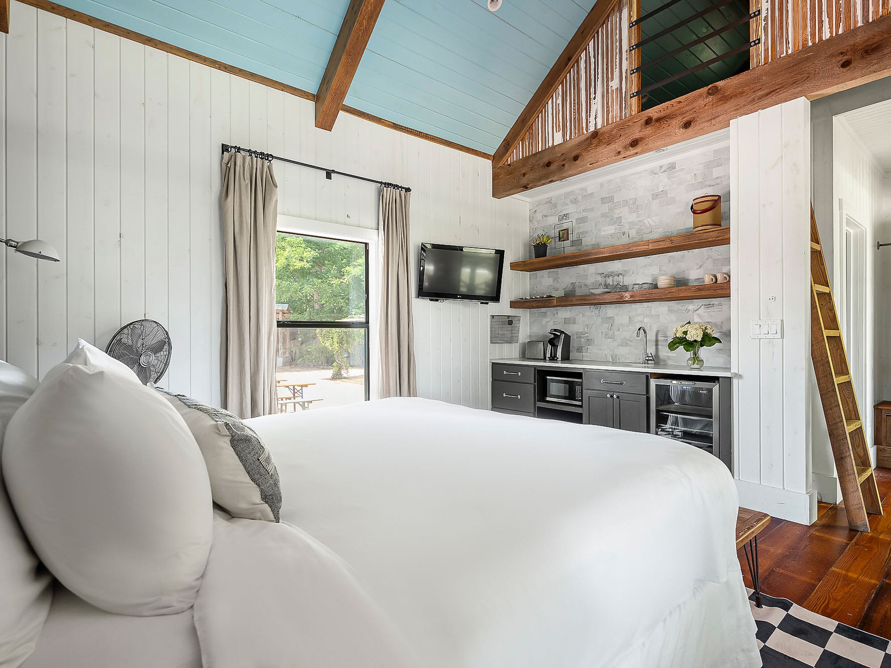 A bedroom with a large bed, a TV mounted on the wall, a kitchenette with a microwave, and a window with curtains.