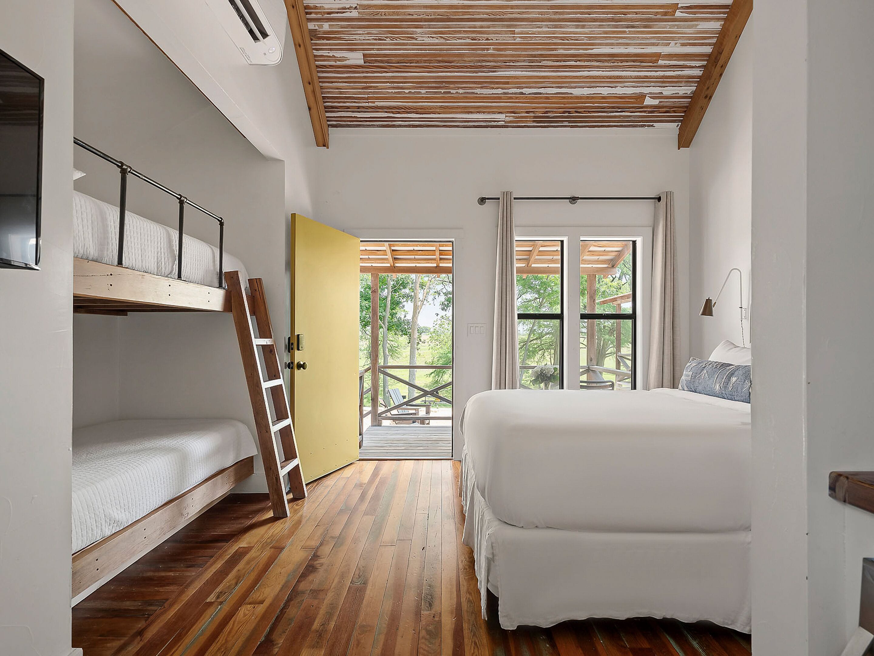 Une chambre avec un lit superposé, un lit simple, un plancher en bois, une porte jaune et une vue sur l’extérieur.