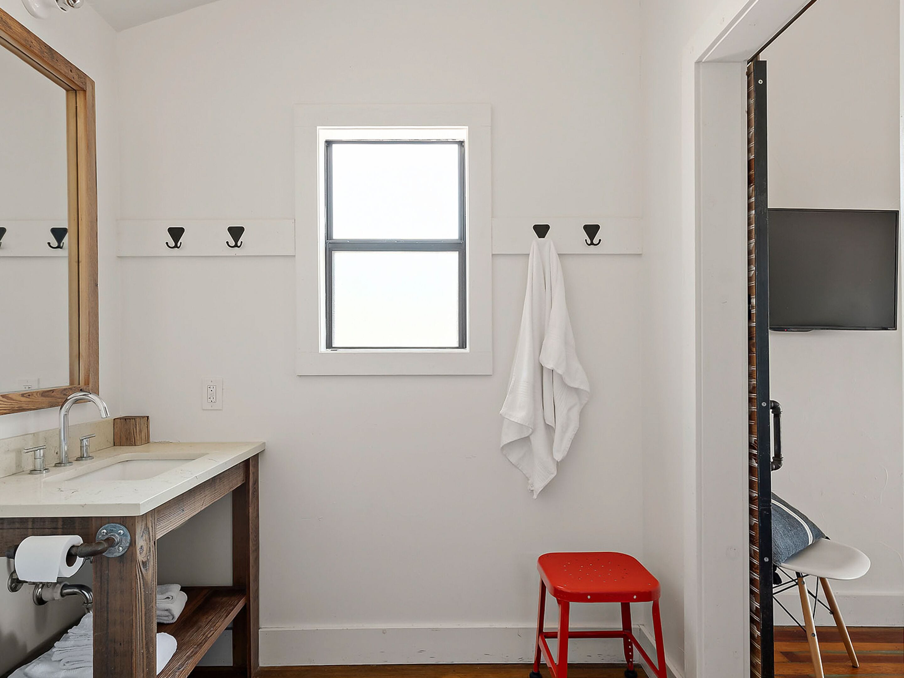 A bathroom with a white sink, wooden vanity, a window, and a red stool.