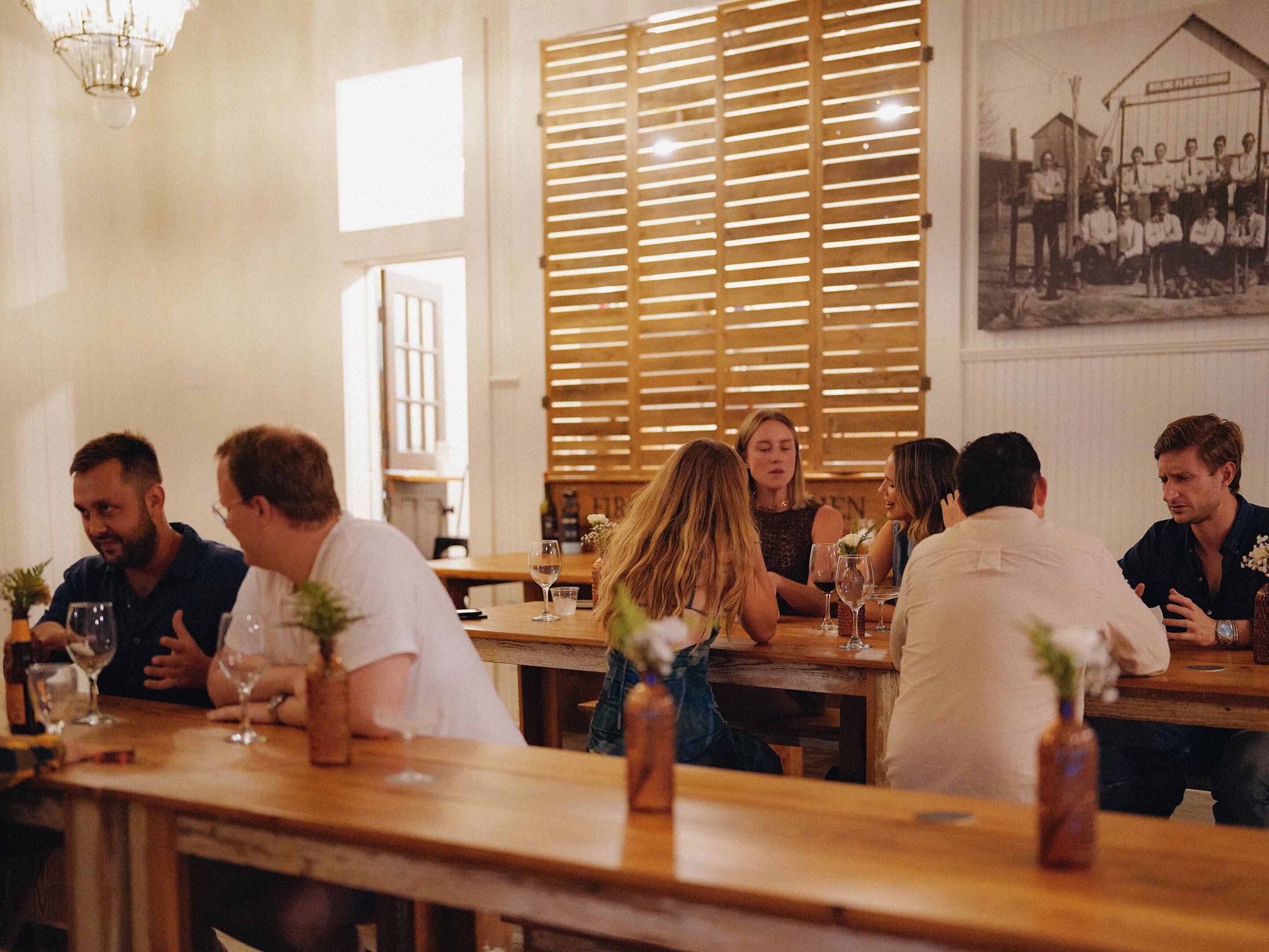 A group of people seated at wooden tables in a restaurant with a large window and wooden slats in the background.