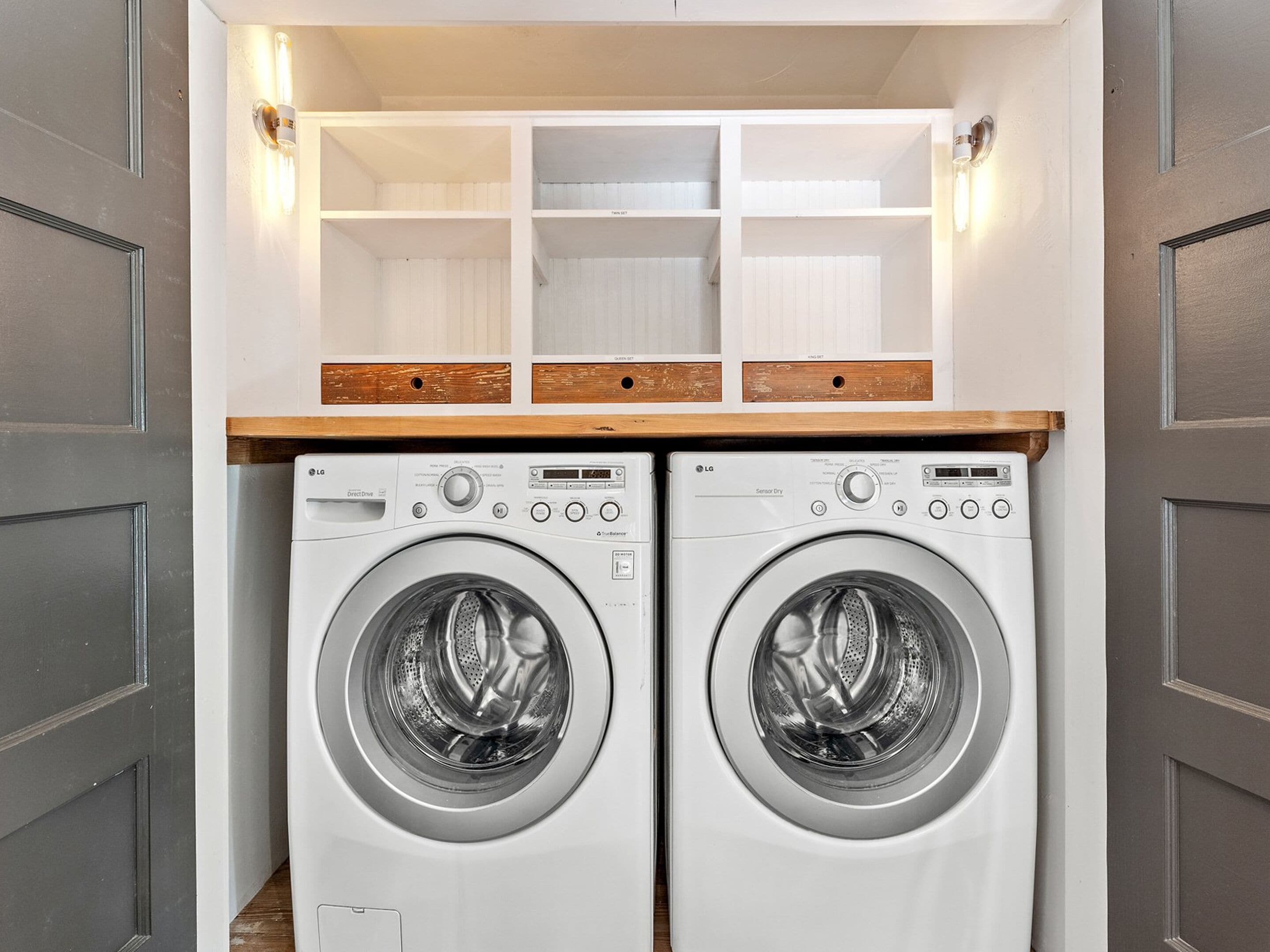 A laundry room with two front-loading washing machines, wooden shelves above, and a white door with a handle.