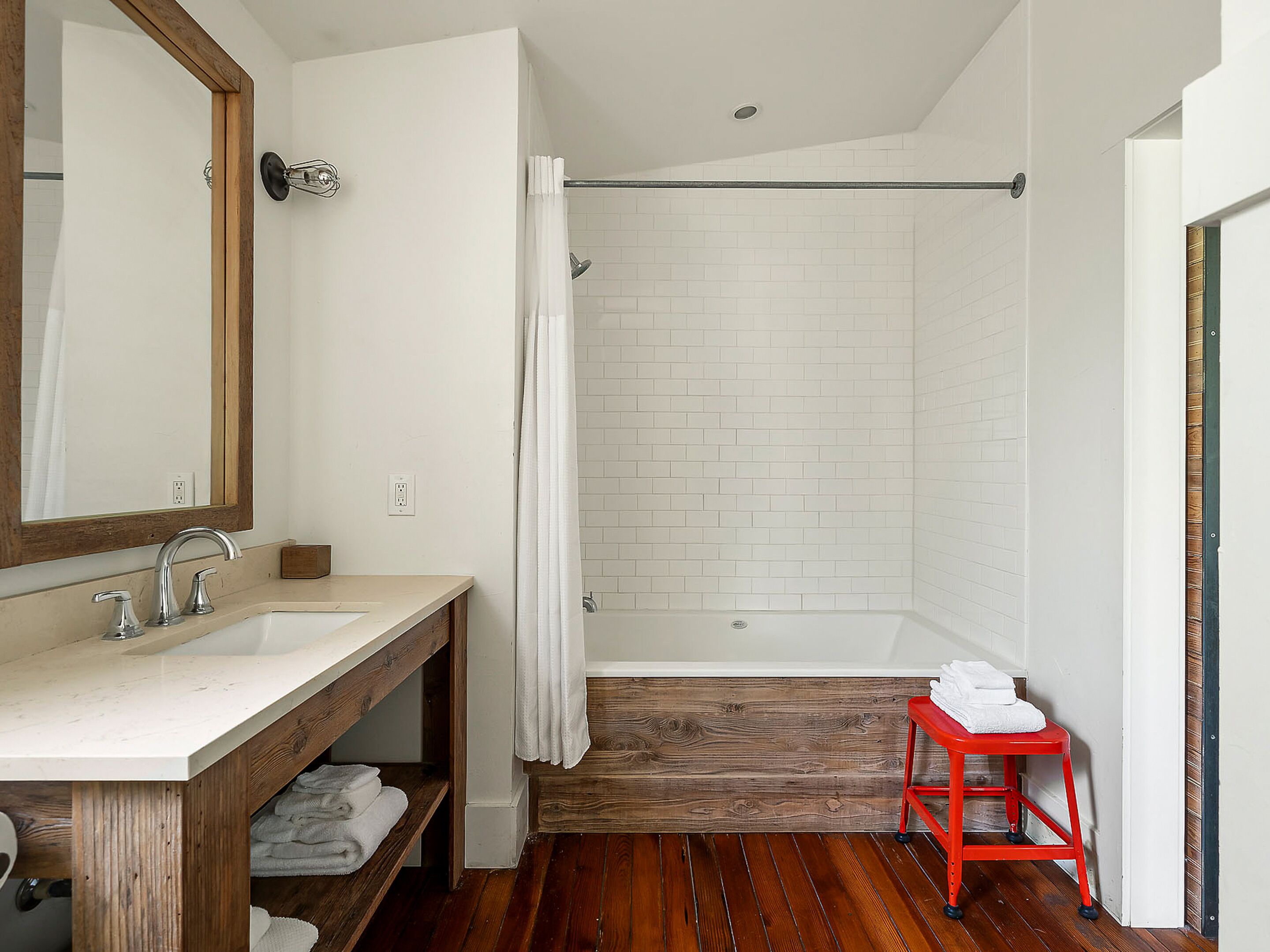 A bathroom with a wooden vanity, a large mirror, a shower with a white curtain, and a red stool.