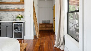 Quarto com teto de madeira, uma janela grande, uma cama, cômoda e área de cozinha com pia e armários.