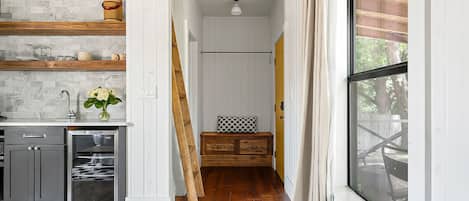 Quarto com teto de madeira, uma janela grande, uma cama, cômoda e área de cozinha com pia e armários.