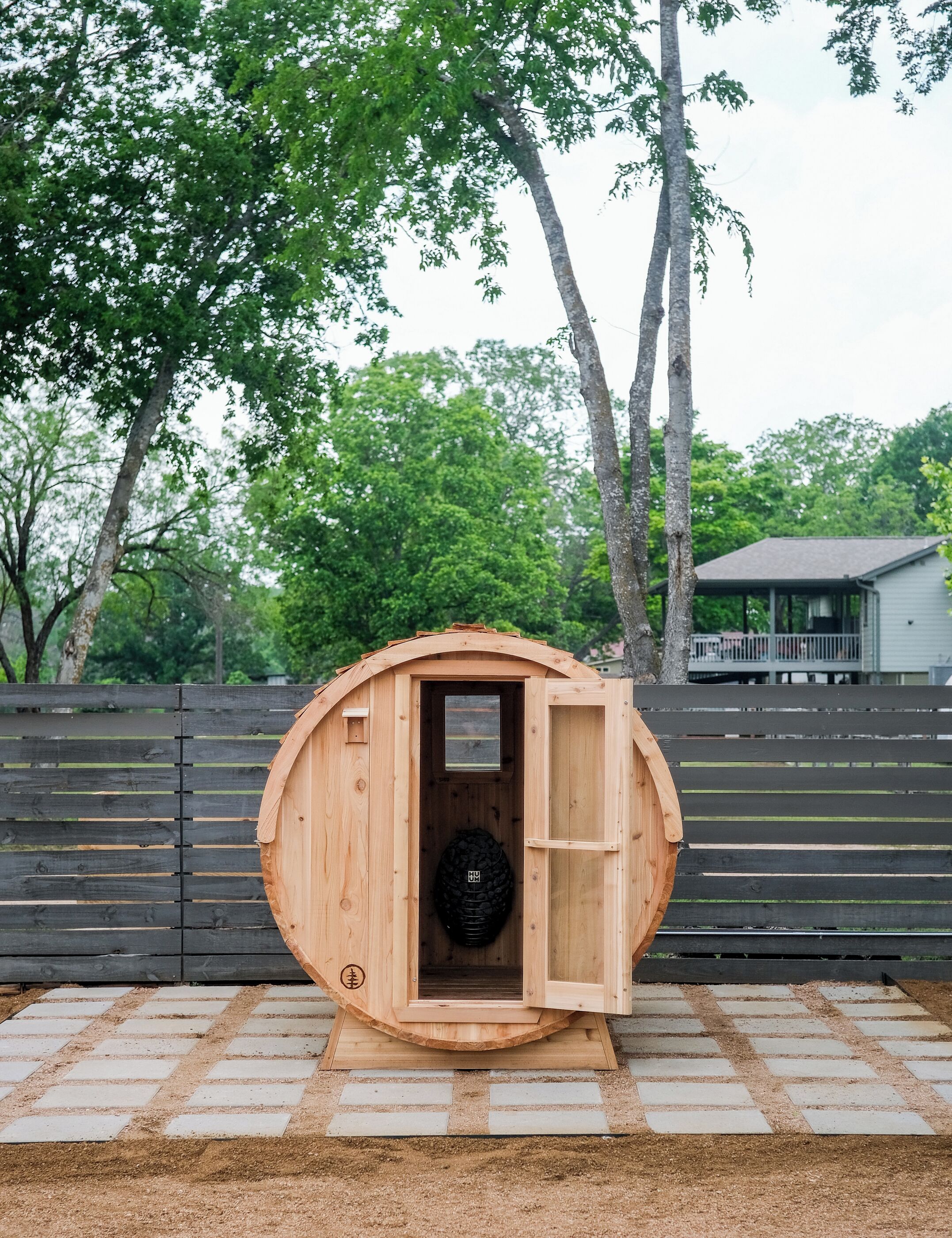 A wooden sauna structure with a door open, a tire in the center, and a paved area in front.