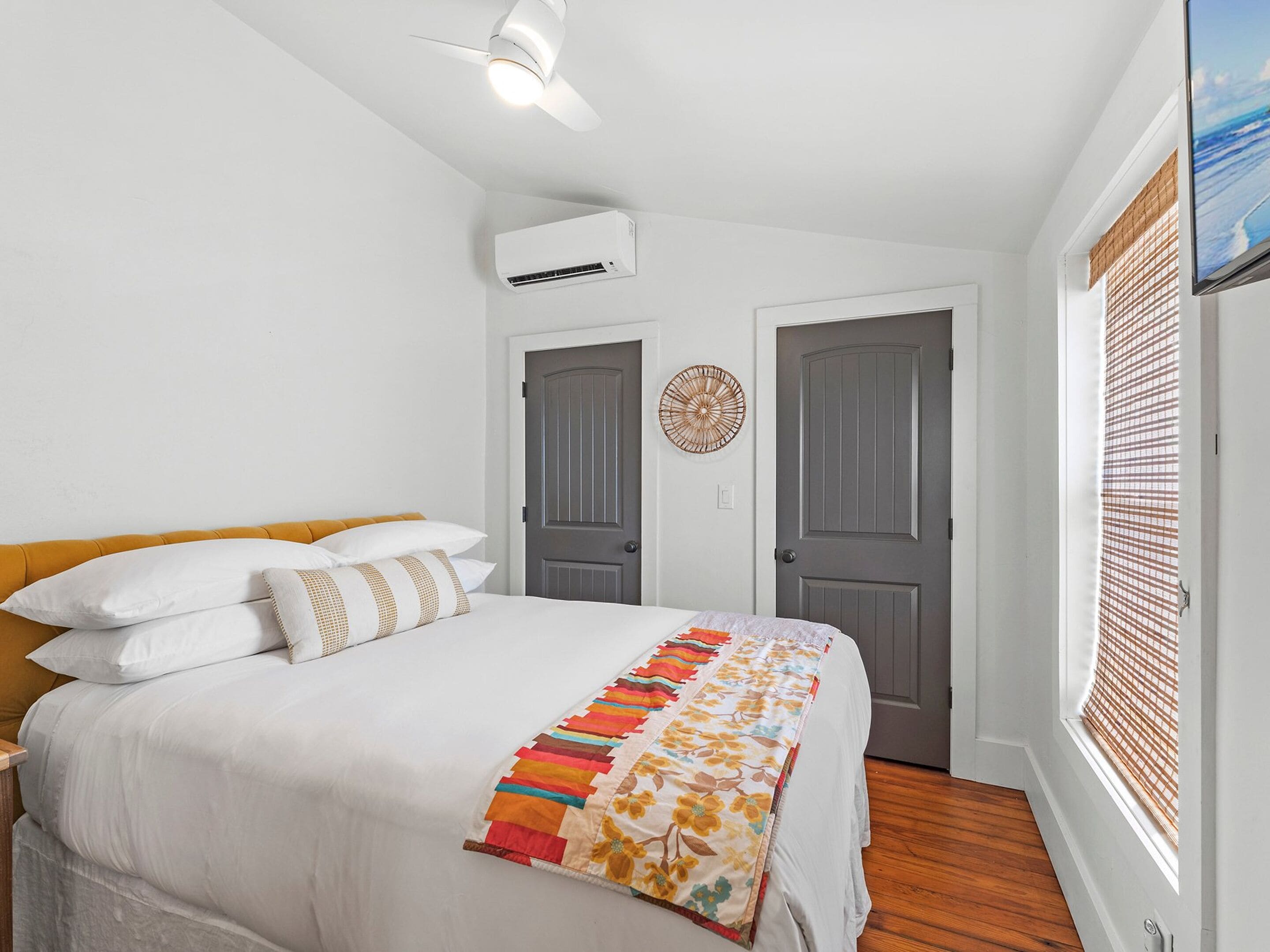 Un dormitorio con una cama, un ventilador de techo, un equipo de aire acondicionado, dos puertas, una ventana con persianas y un adorno en la pared.
