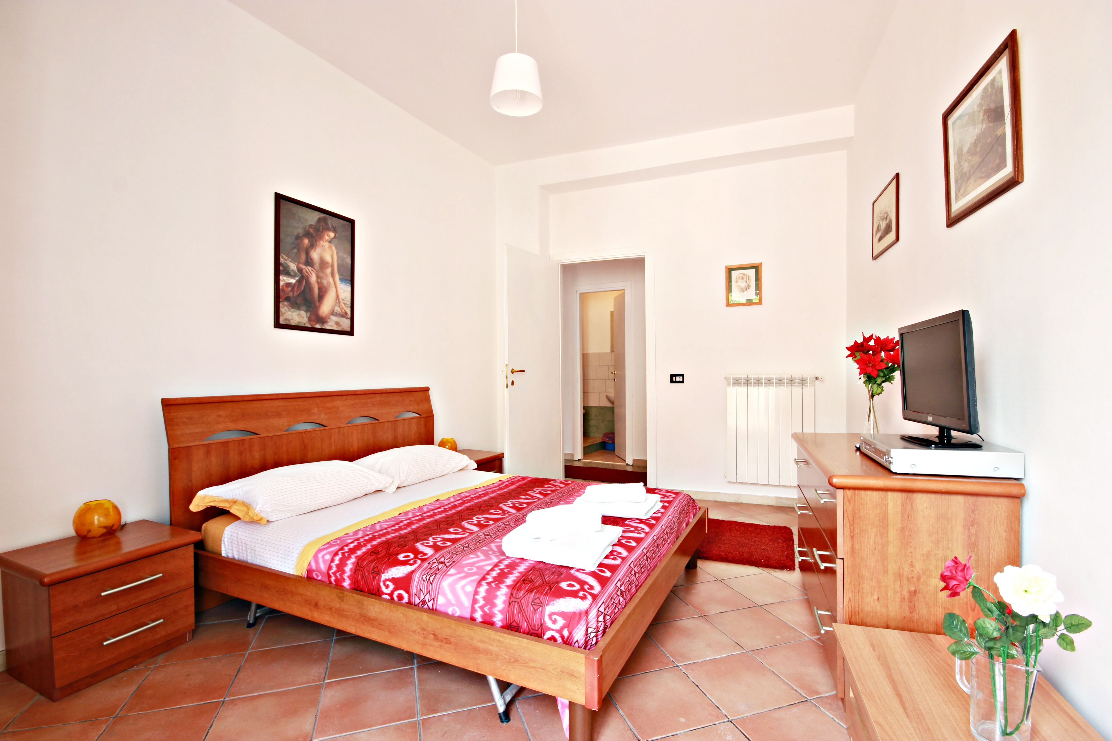 A bedroom with a wooden bed, a nightstand, a desk with a computer, and a TV.