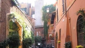 A narrow cobblestone street flanked by buildings with arched doorways and windows, some with plants. There are outdoor seating areas and a few parked vehicles. - Stairs of Trastevere (Rome)