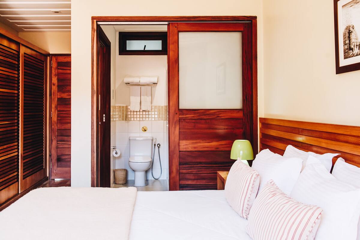 A bedroom with a wooden headboard, a bed with white bedding, a nightstand with a green lamp, and a view into the bathroom with a toilet and shower.