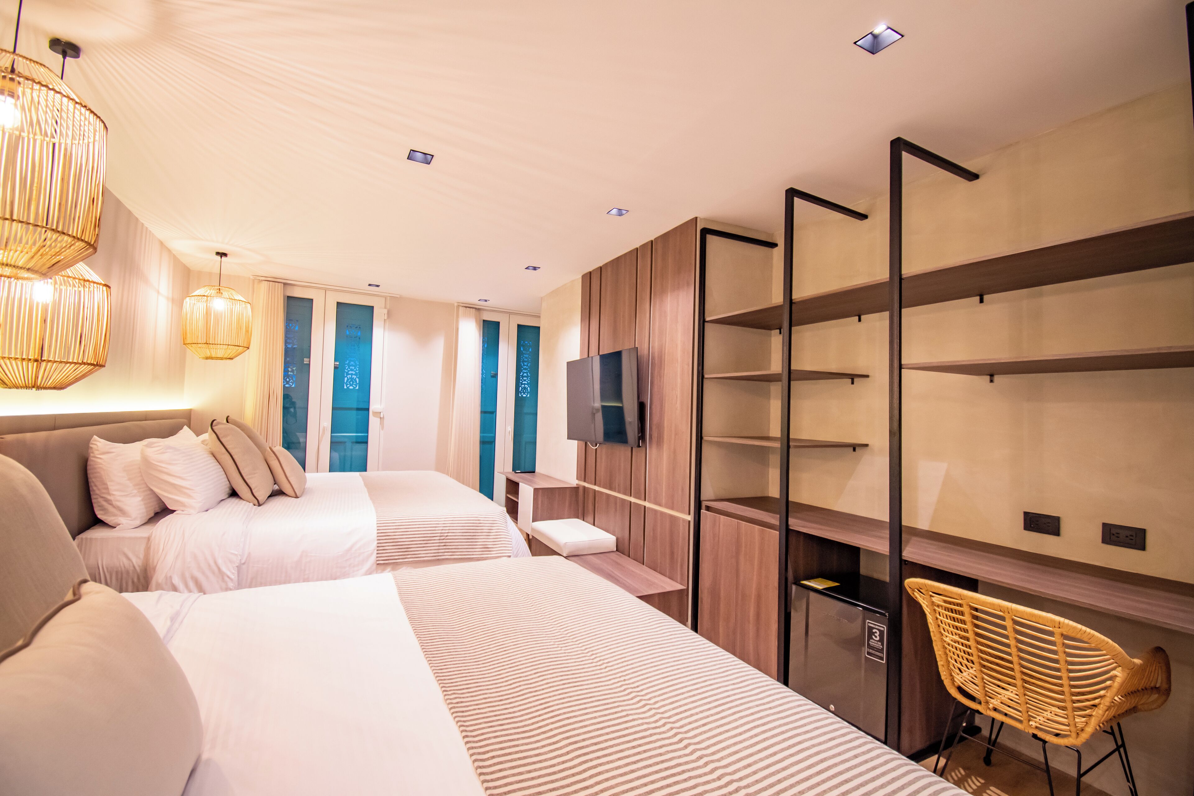 A modern hotel room with two beds, a TV, a wooden wardrobe, and a wicker chair.