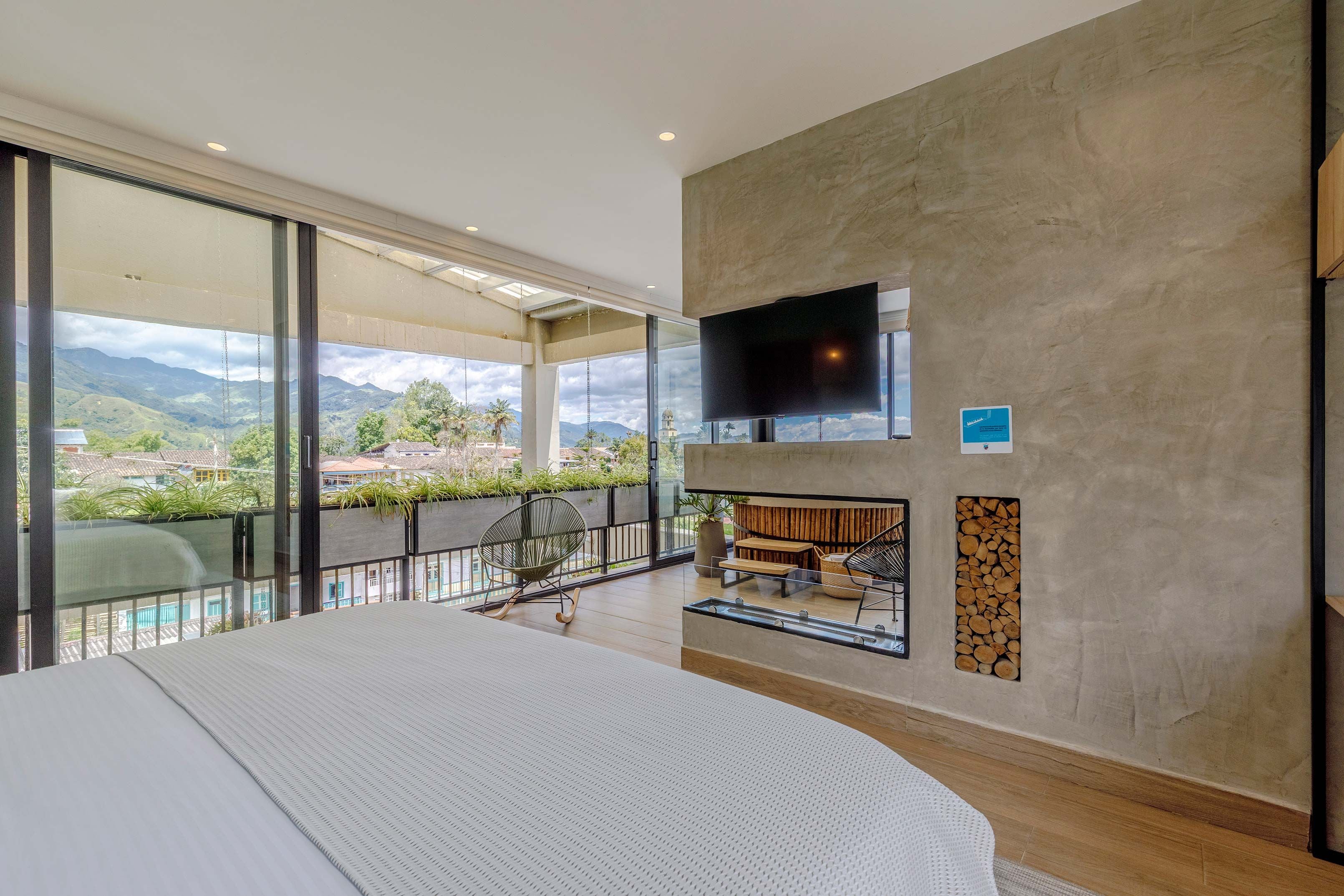 A modern hotel room with a large bed, a TV mounted on the wall, a fireplace, and a balcony with a view of the mountains.