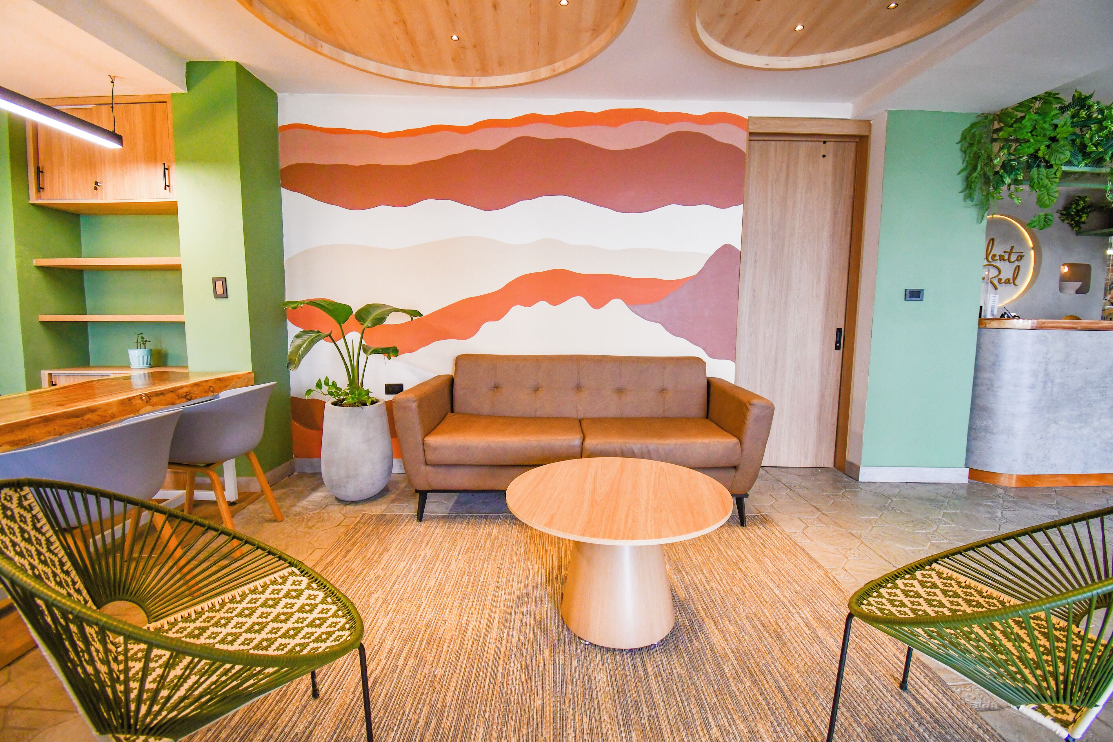 Un área de estar moderna con un sofá, dos sillas verdes, una mesa de centro de madera, un mural con formas abstractas y un mostrador de recepción con una planta.